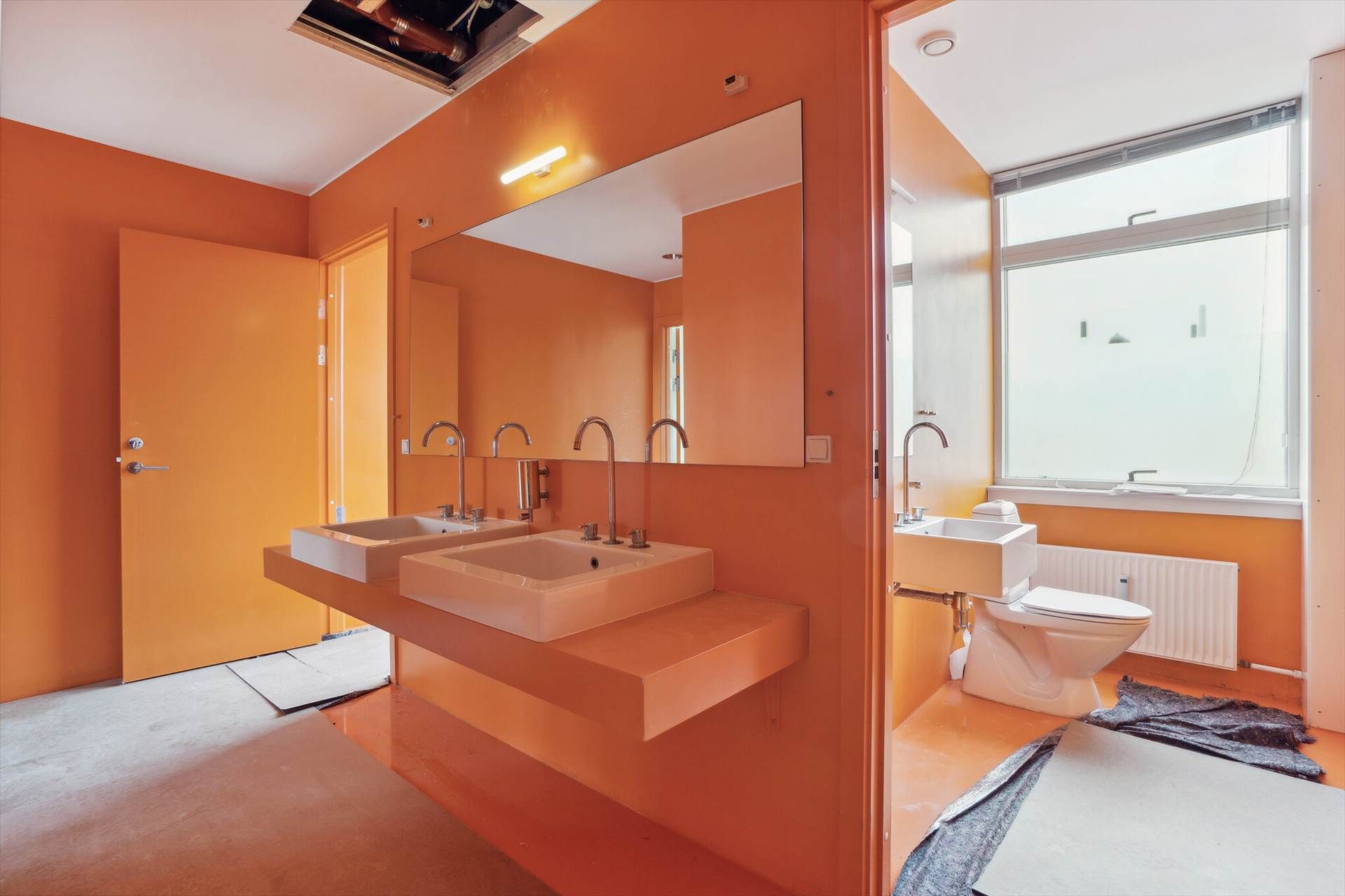 Et lyst badeværelse med orange vægge, lyse gulve og to store vasker med hver deres vandhane samt et toilet og en radiator