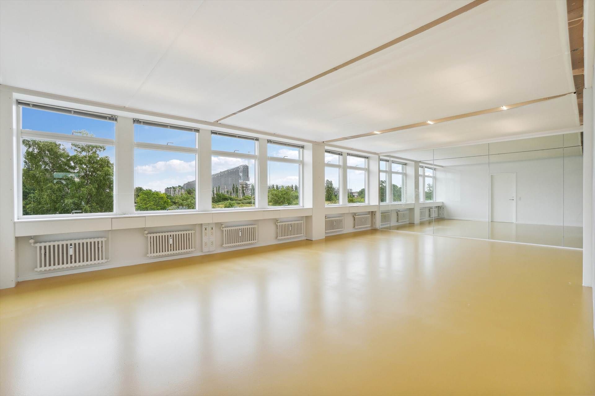 Et stort, ubenyttet kontor med store vinduer og spejl. Gulvet er hvidt og smedet, og der er flere radiatorer.