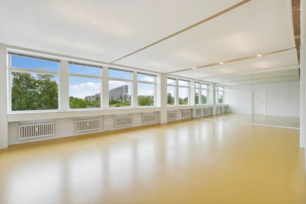 Et stort, ubenyttet kontor med store vinduer og spejl. Gulvet er hvidt og smedet, og der er flere radiatorer.