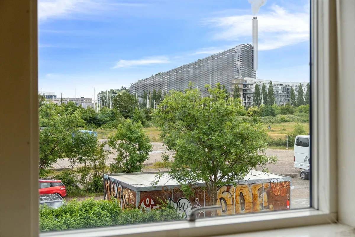 Et stort byggeri i bakkeform med en røgsøjle i baggrunden, og en stor container med graffiti og en hvid bus i forgrunden