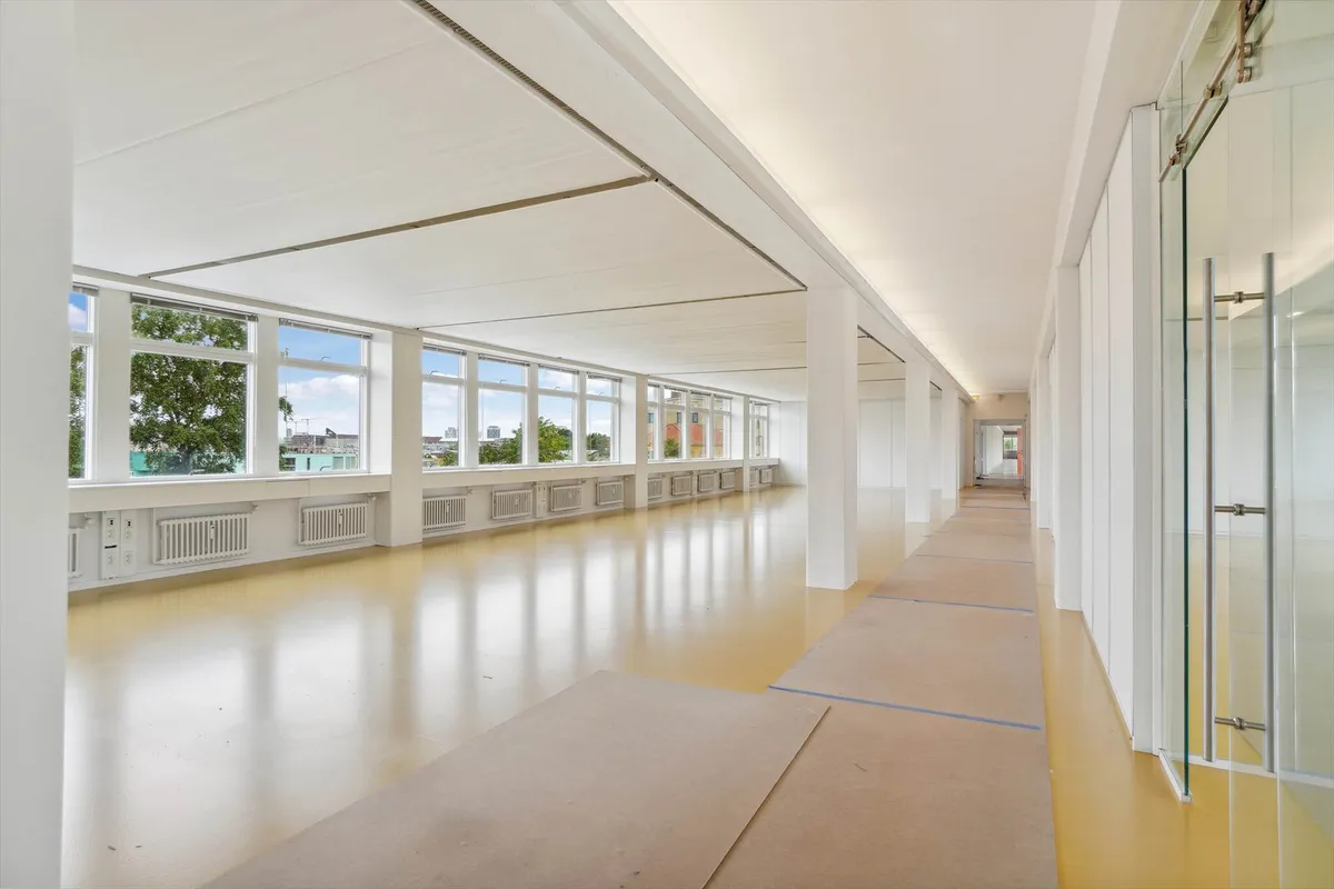 To store kontorlokaler med hvide vægge, gulv og loft, store vinduer, gulgulvt og hvide søjler, tavler og kontakter på væggen