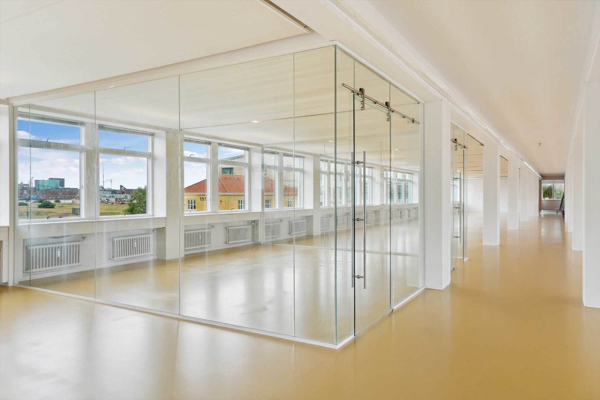 Et moderne kontor med glasvægge og højloft, hvor vinduer giver udsigt til bygninger og grønne områder udenfor