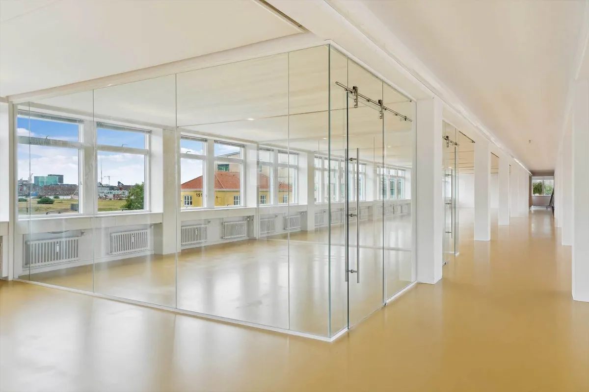 Et moderne kontor med glasvægge og højloft, hvor vinduer giver udsigt til bygninger og grønne områder udenfor