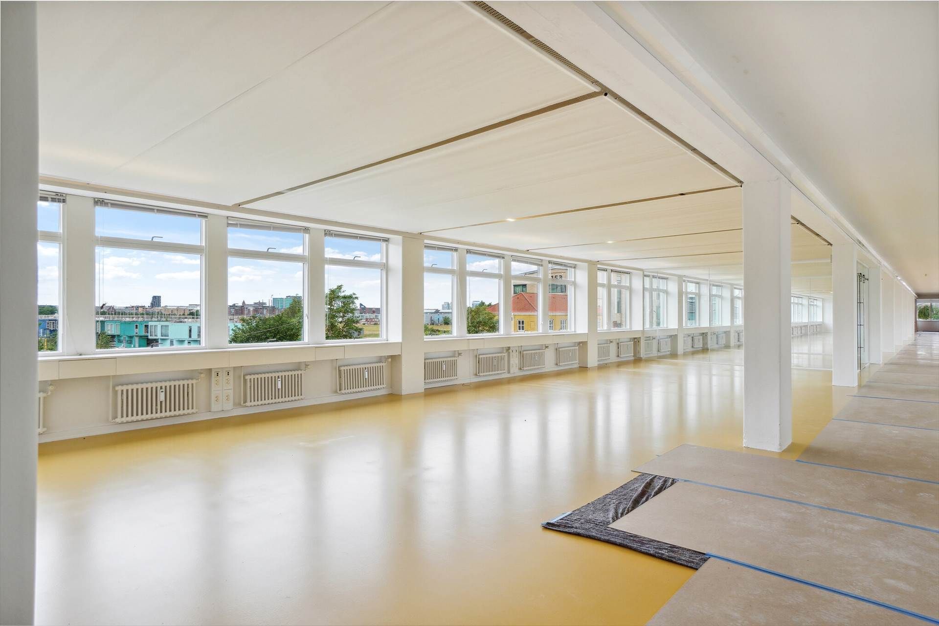 Et rummeligt kontor med hvide vægge og gulve, højt til loftet, store vinduer, og trægulv på den ene side