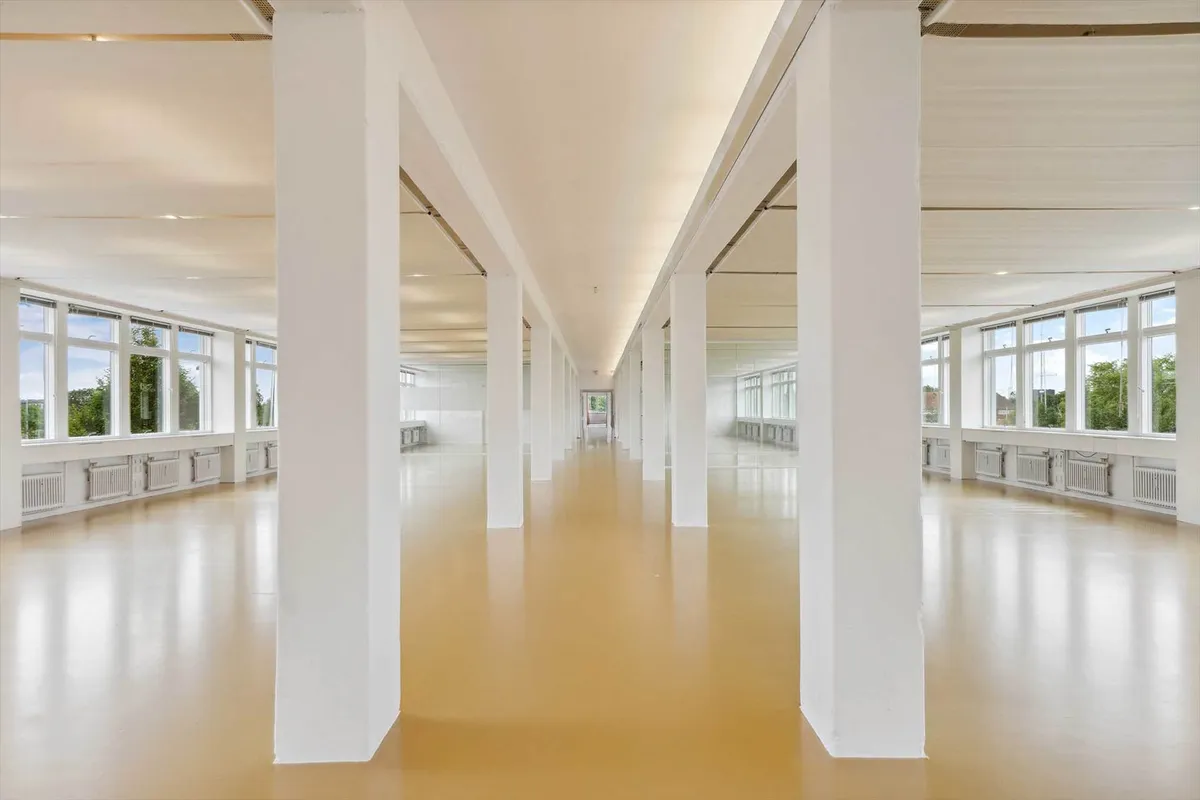 Lyst og rummeligt kontor med højloft og lyse gulve og vinduer, der giver god udsigt til udendørsarealer