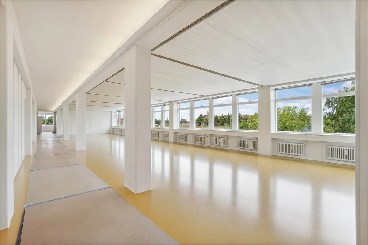 Et lyst og rummeligt kontor med hvide vægge, gulve og loft, samt store vinduer med udsigt til træer og bygninger