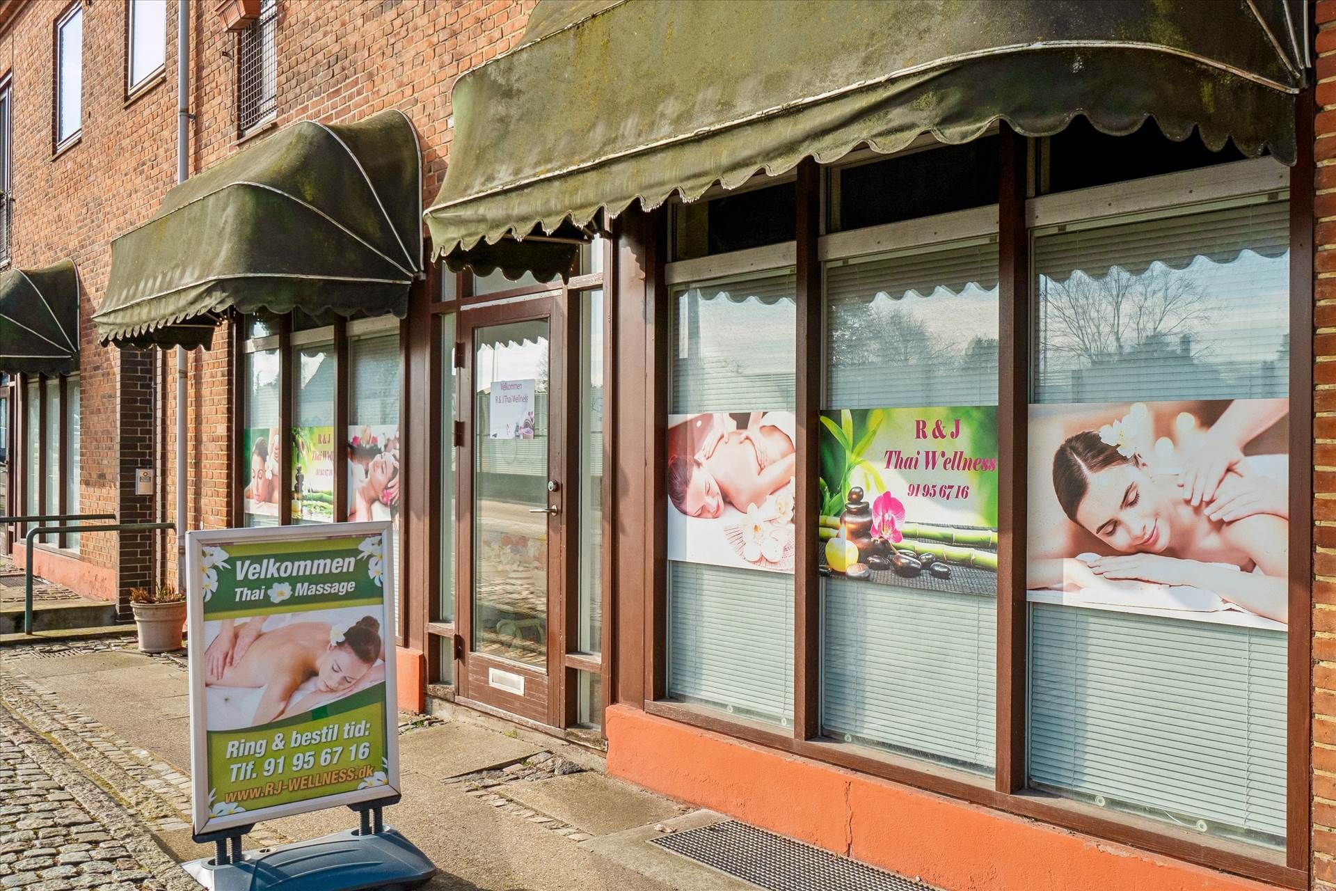 En butik med tre vinduer og reklameplakater med massagereklamer og åbningstider. Butikken har tre grønne tagskærme og en vej