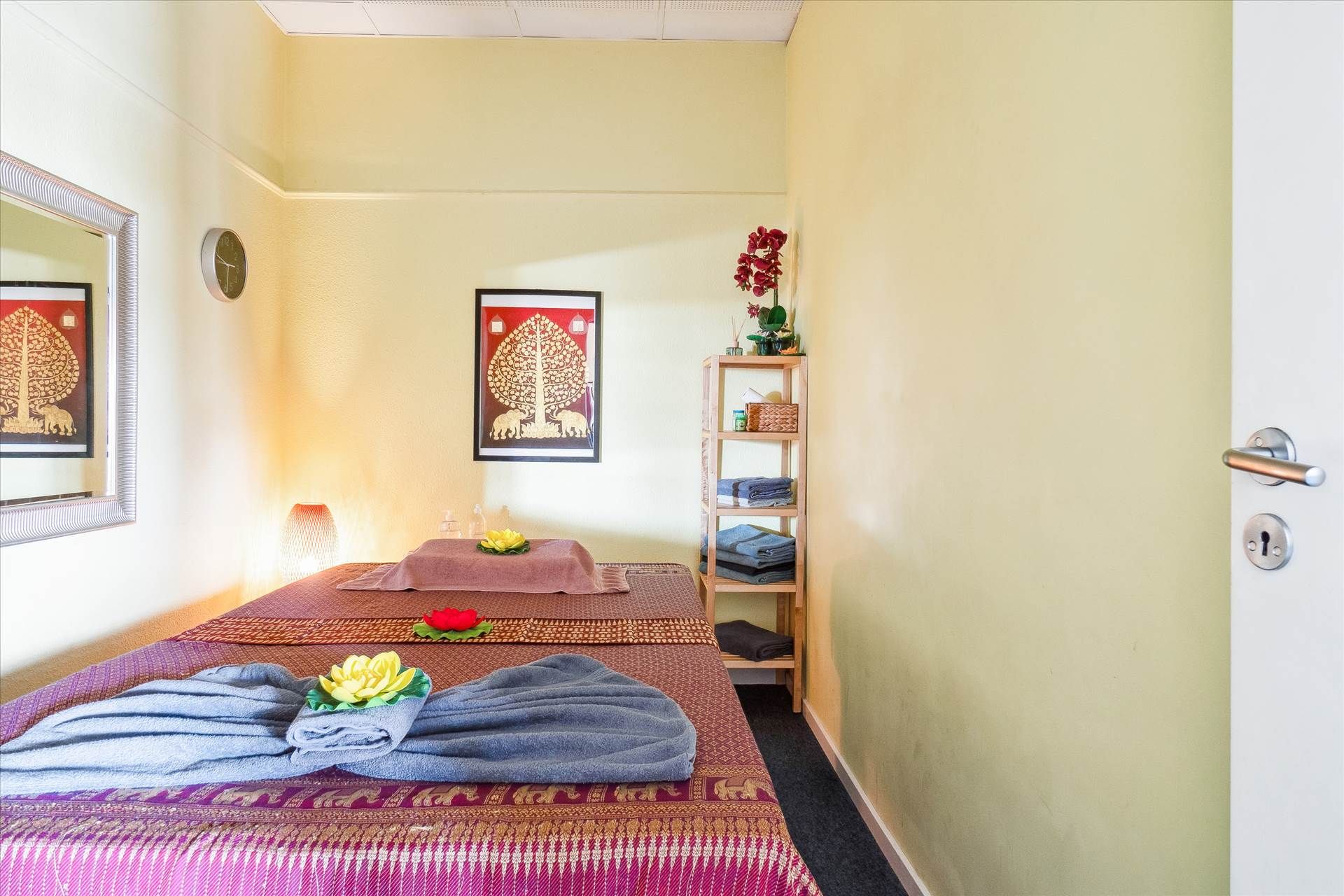 Massørrum med blomster og to massagebord dækket med lyserøde håndklæder og tøj samt gulvlampe og kunstværk på væggen