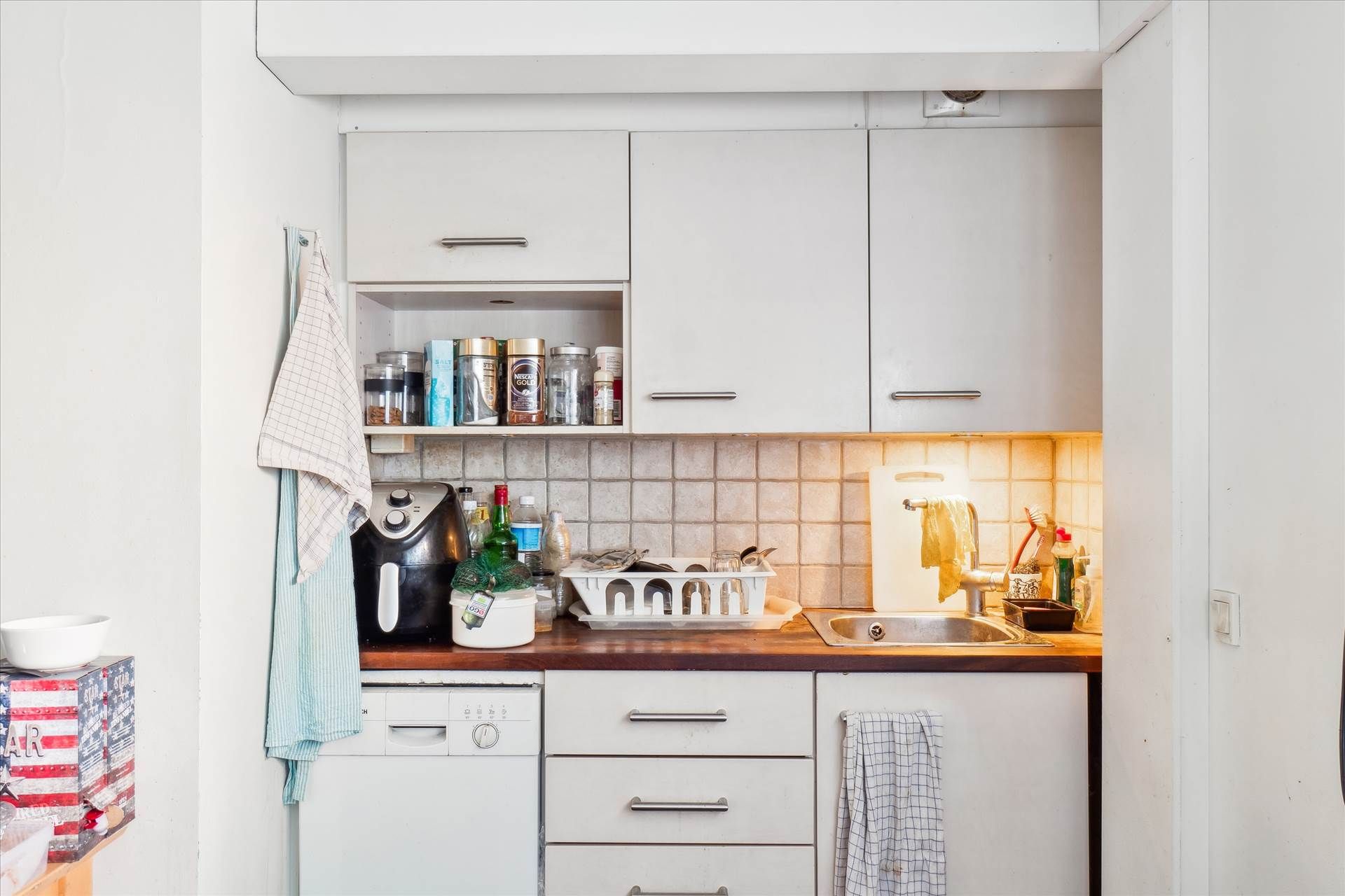 Køkken med hvide vægge, hvide køkkenskabe og disk, hvor der er en hvid skuffe med en stang og en klud hængende på den