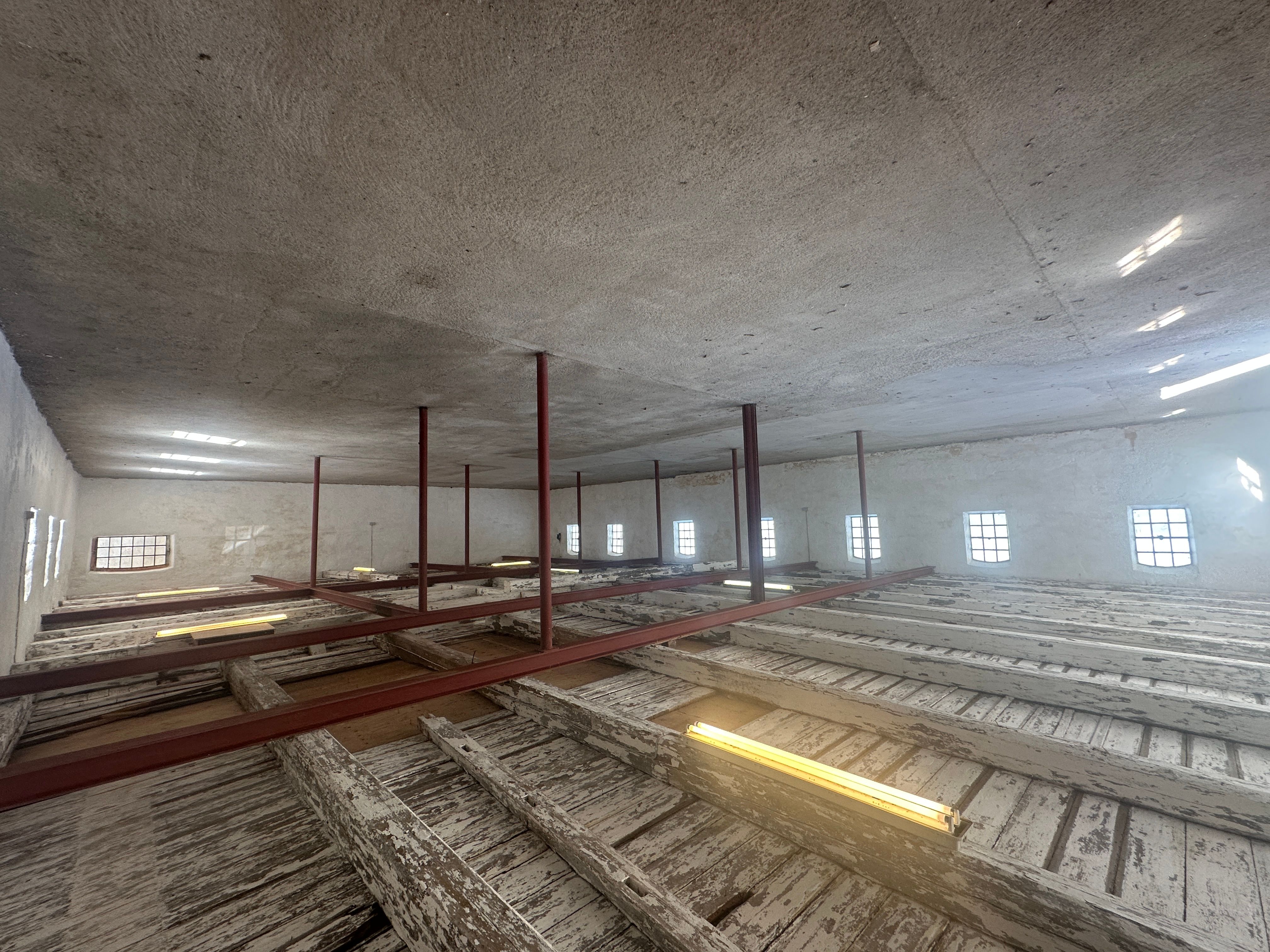 Lagerrum med store vinduer og rå hvide vægge og gulv. Store røde stålbjælker under loftet. Mange brædder står på gulvet.