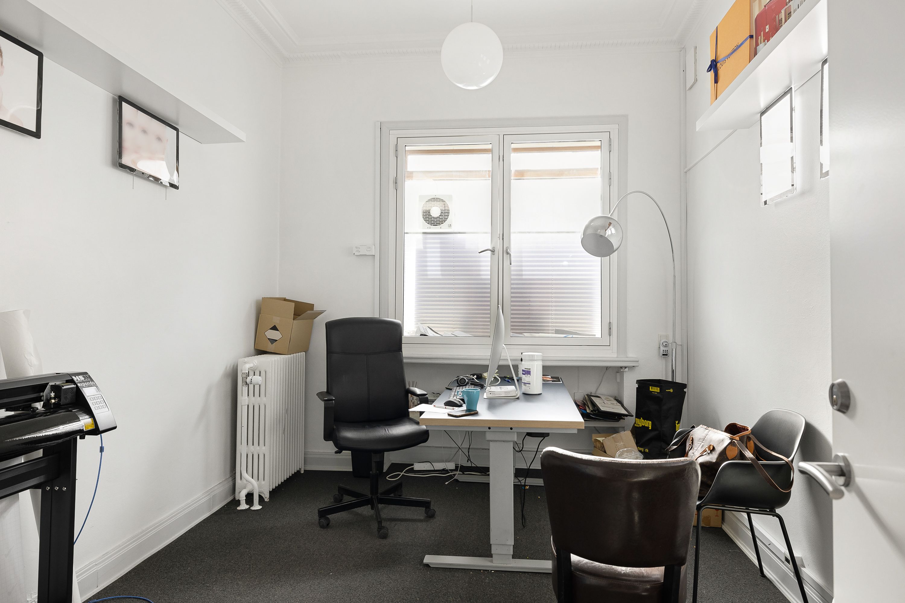 Et kontorlokale med en høj skrivebord, stålstol, kaffemaskine og en lampeskærm på gulvet og en væg med et skrivebord, en