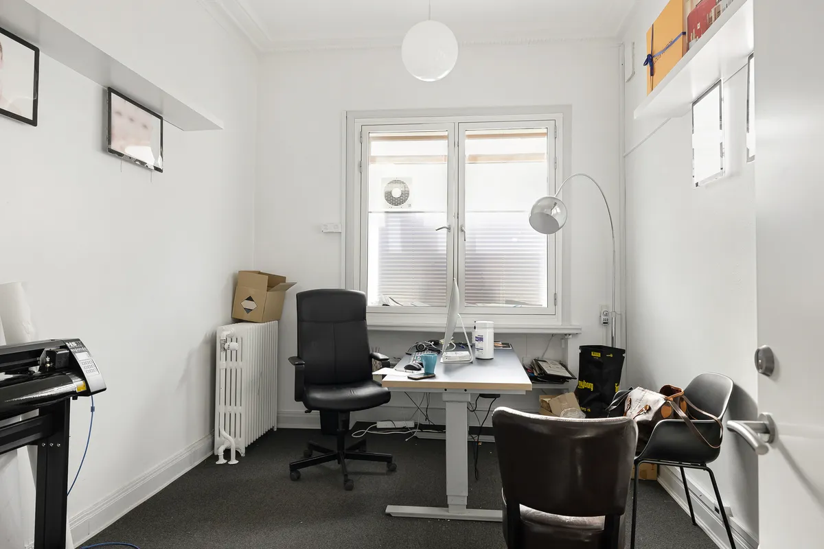 Et kontorlokale med en høj skrivebord, stålstol, kaffemaskine og en lampeskærm på gulvet og en væg med et skrivebord, en