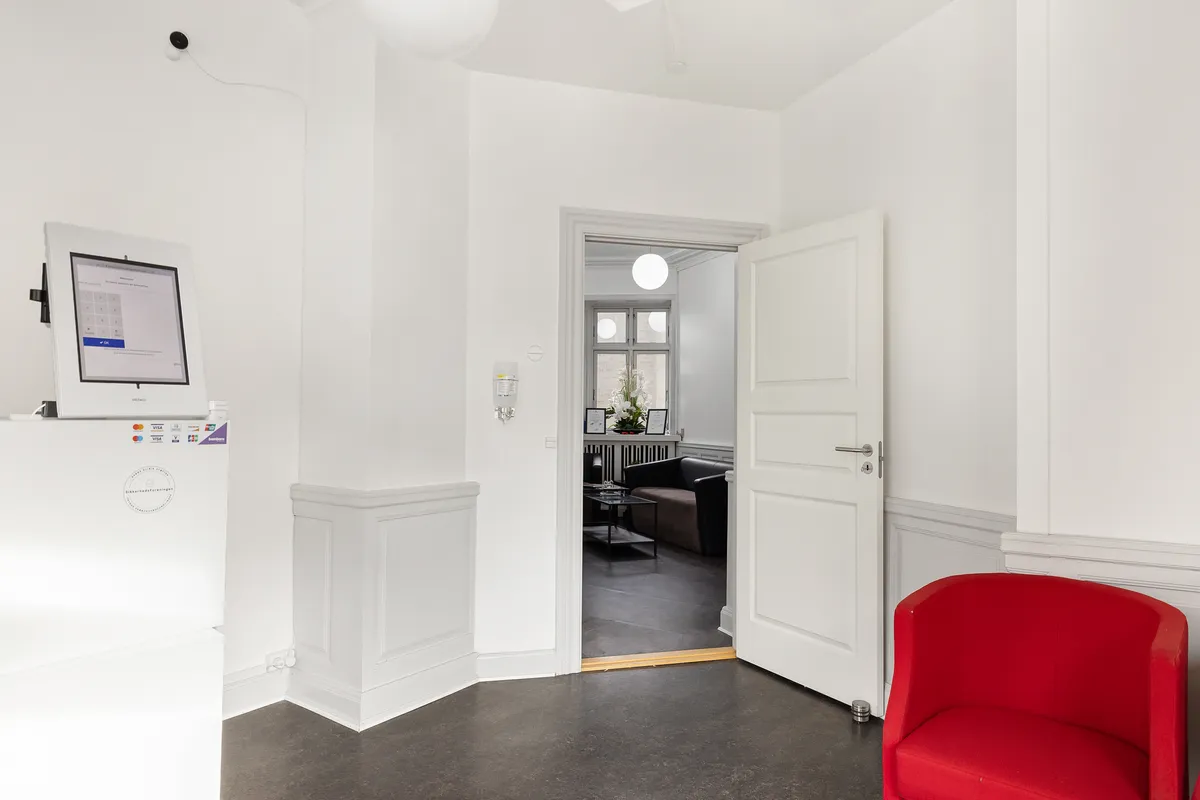 Kontorområde med en hvid væg, en rød stol, en åben dør og et skrivebord med en monitor og et køleskab med kreditkort og et