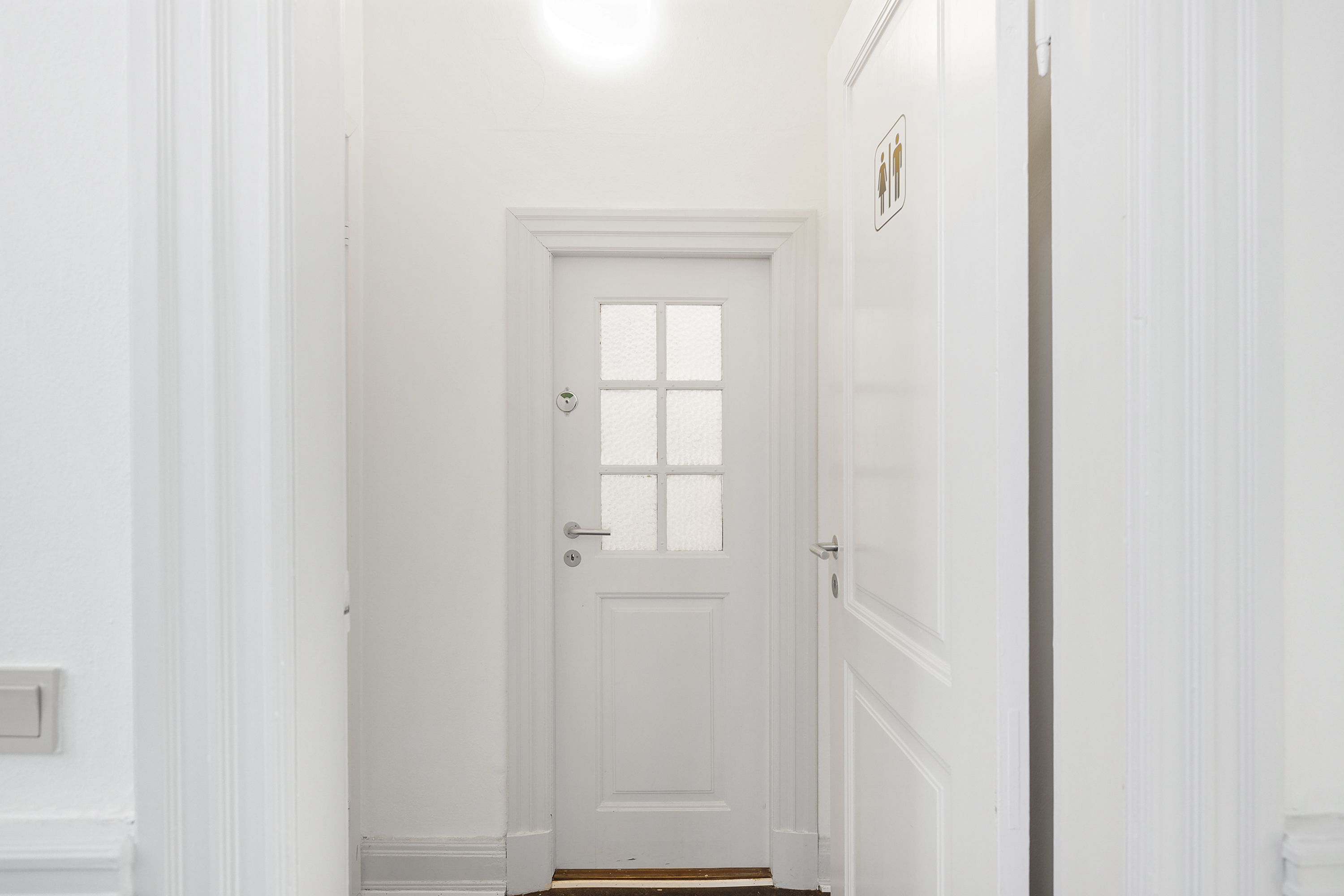En hvid hall med to hvide døre og en hvid væg med et toiletskilt på den ene side af døren og en åben dør på den anden side