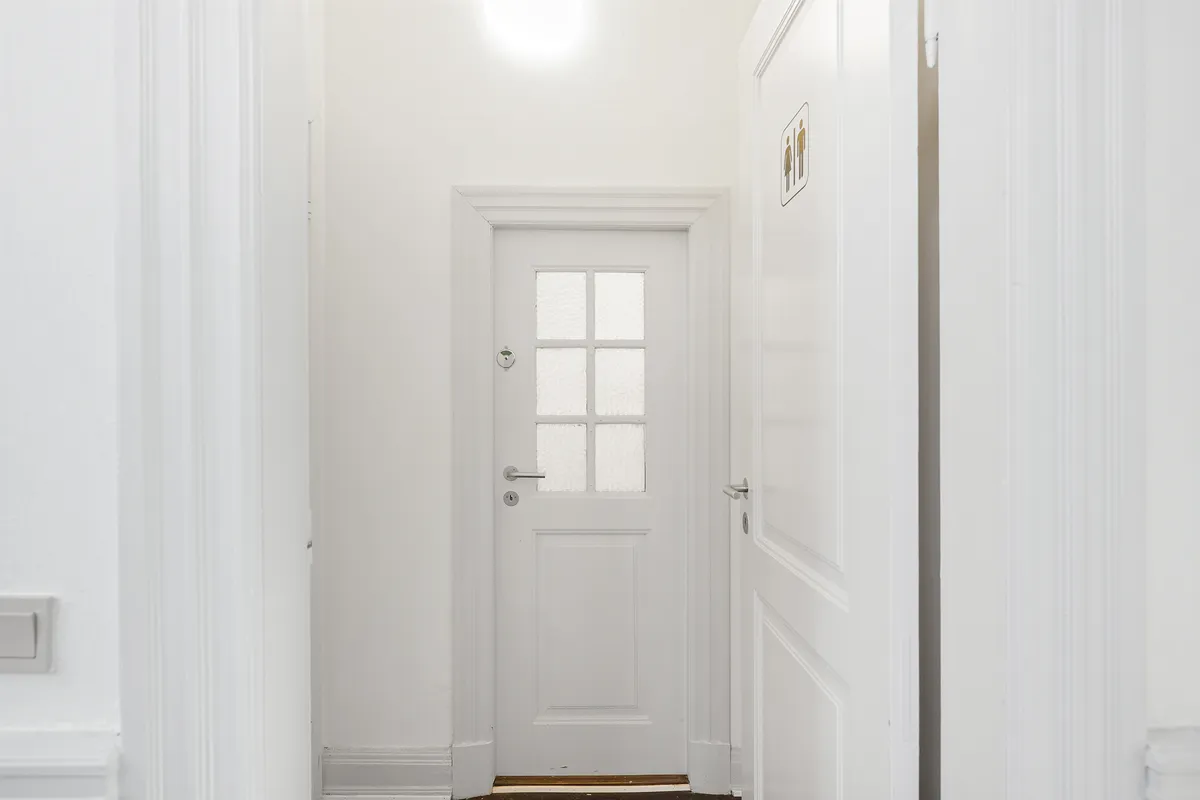 En hvid hall med to hvide døre og en hvid væg med et toiletskilt på den ene side af døren og en åben dør på den anden side