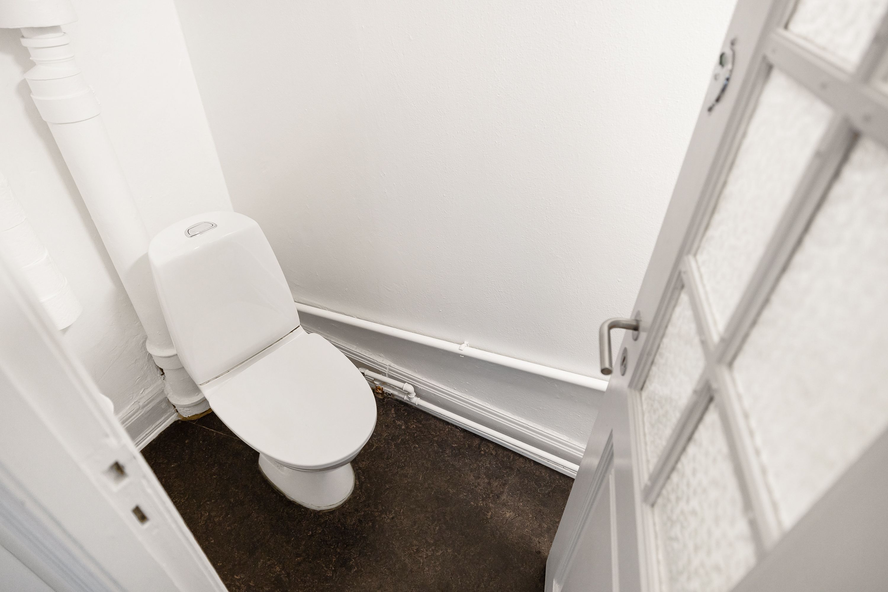 Rent, hvidt toilet med papirhylde og åben dør, der fører ud til korridor, gulv og væg i lyst hvidt