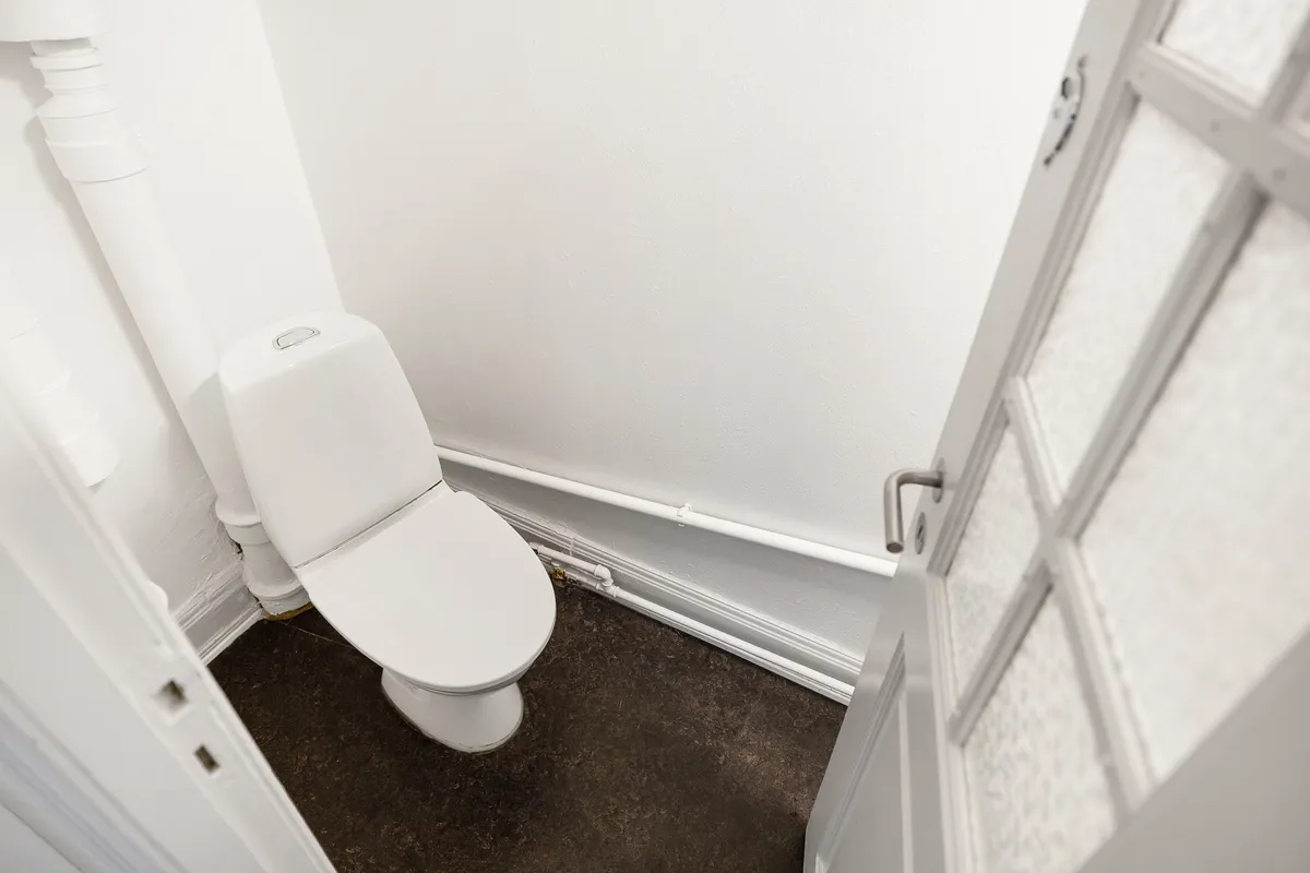 Rent, hvidt toilet med papirhylde og åben dør, der fører ud til korridor, gulv og væg i lyst hvidt