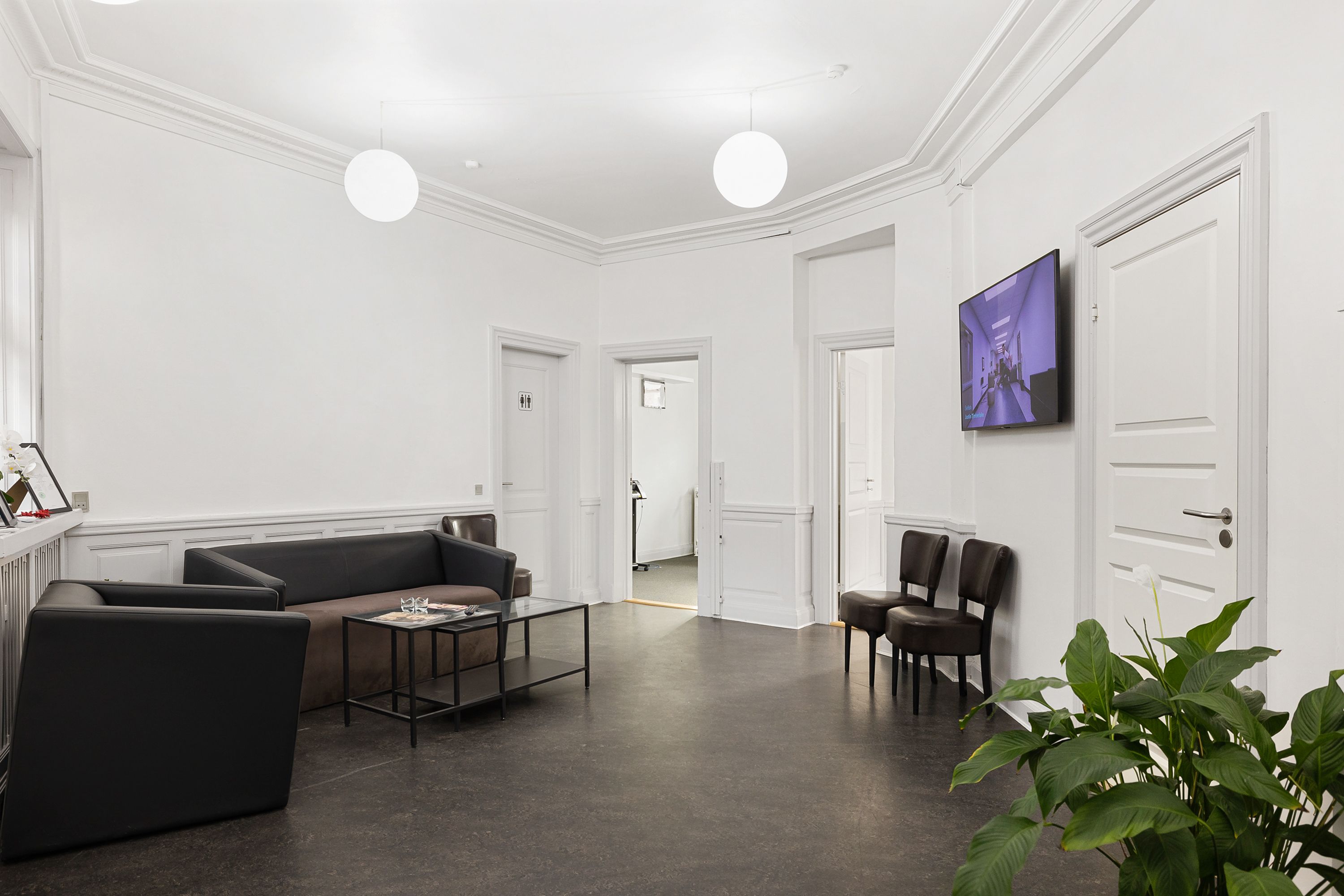 Minimalistisk indretning med hvide vægge, mørke sofaer, to stole, en glasbord, en tv-skærm og et gulv med jordfarve