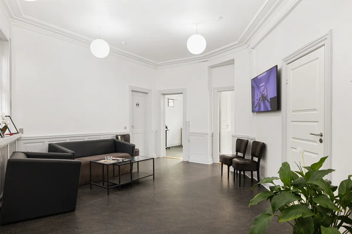 Minimalistisk indretning med hvide vægge, mørke sofaer, to stole, en glasbord, en tv-skærm og et gulv med jordfarve