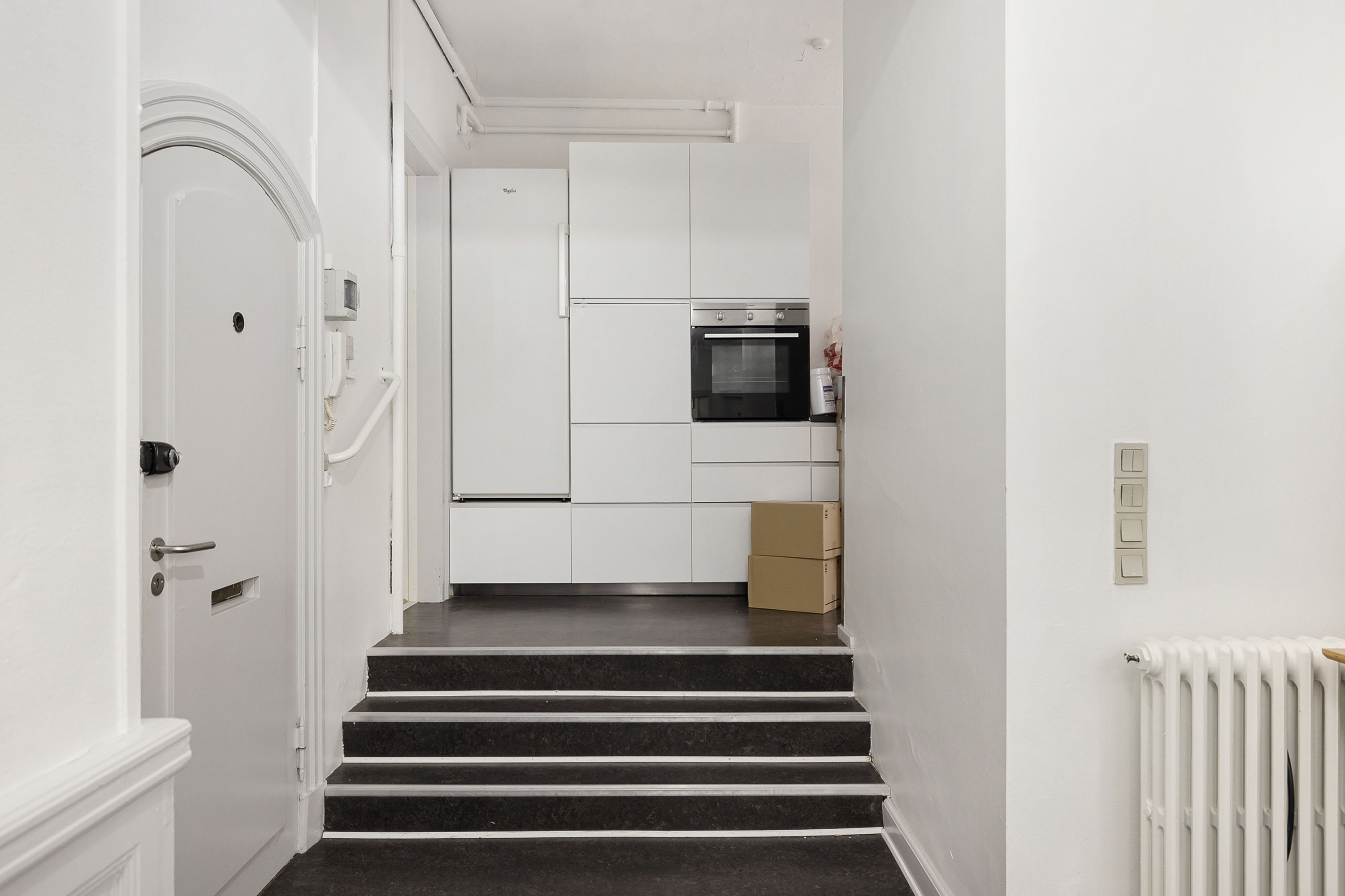 En mørk trappe fører op til en hvid køleskab og indbygget ovn i en stilren gang. Højre er der en radiator og tre tænd/sluk