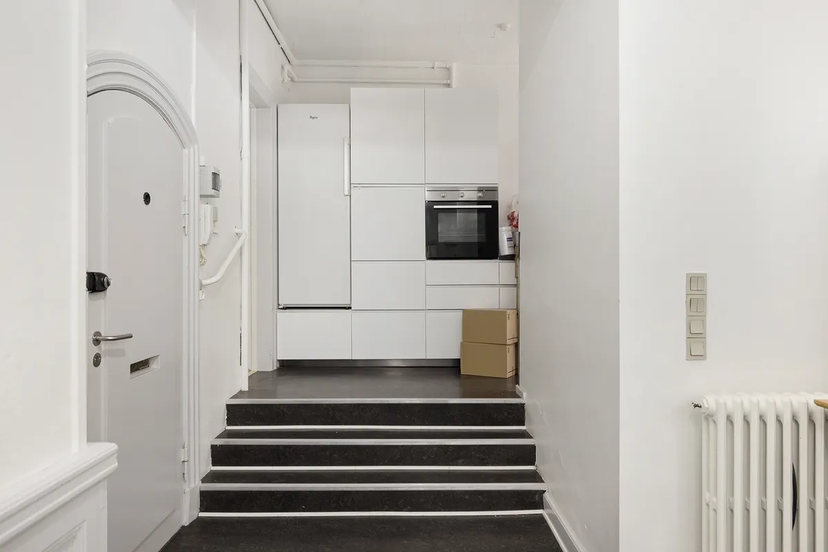 En mørk trappe fører op til en hvid køleskab og indbygget ovn i en stilren gang. Højre er der en radiator og tre tænd/sluk