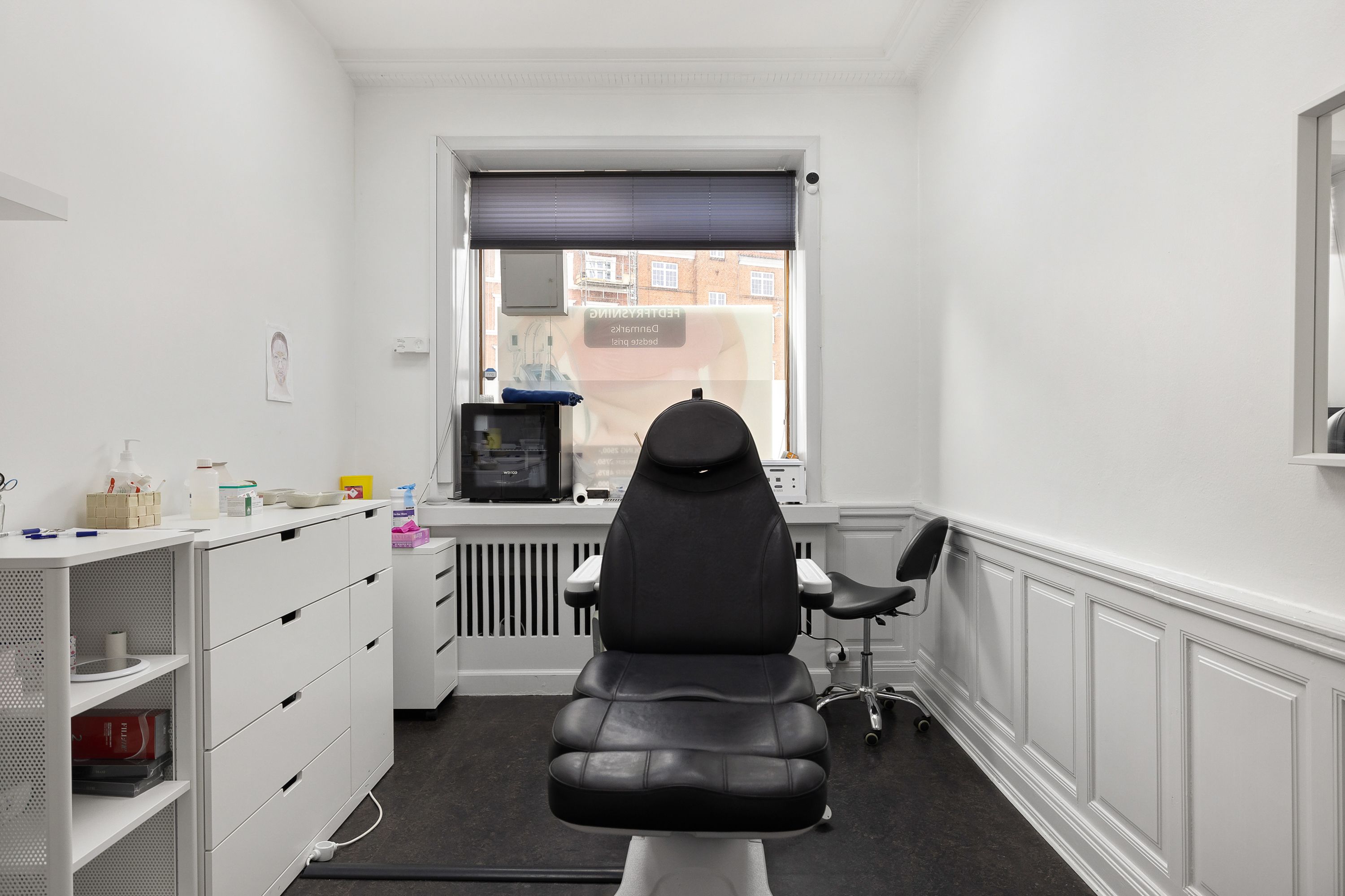 En tandlægerstol og en hjælpestol står på en sort gulvbelægning i en hvid vægget og gulvvæggede tandlægeklinik