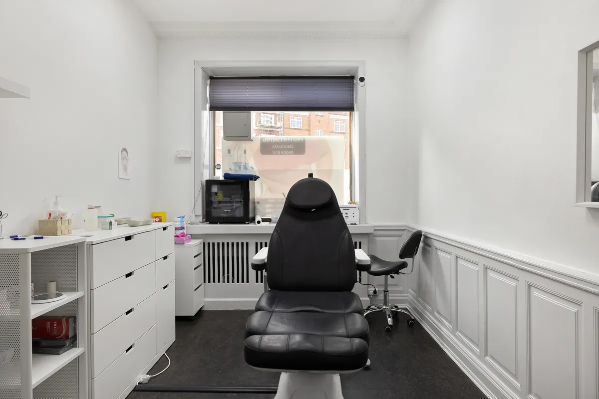 En tandlægerstol og en hjælpestol står på en sort gulvbelægning i en hvid vægget og gulvvæggede tandlægeklinik