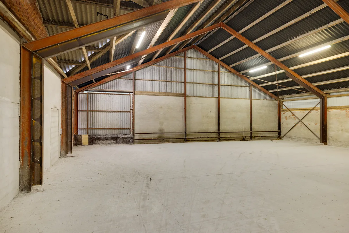 En stor lagerhal med metalliske loftsbjælker og vægge. Der er en åben dør på højre side. Lyspærer er fastgjort til loftet.