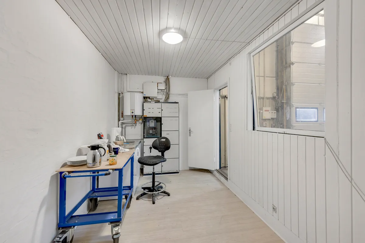 Hjørne af et moderne køkken med hvide vægge og lysekroner, rullebord med kaffekande og en højstole samt dør til en anden rum.