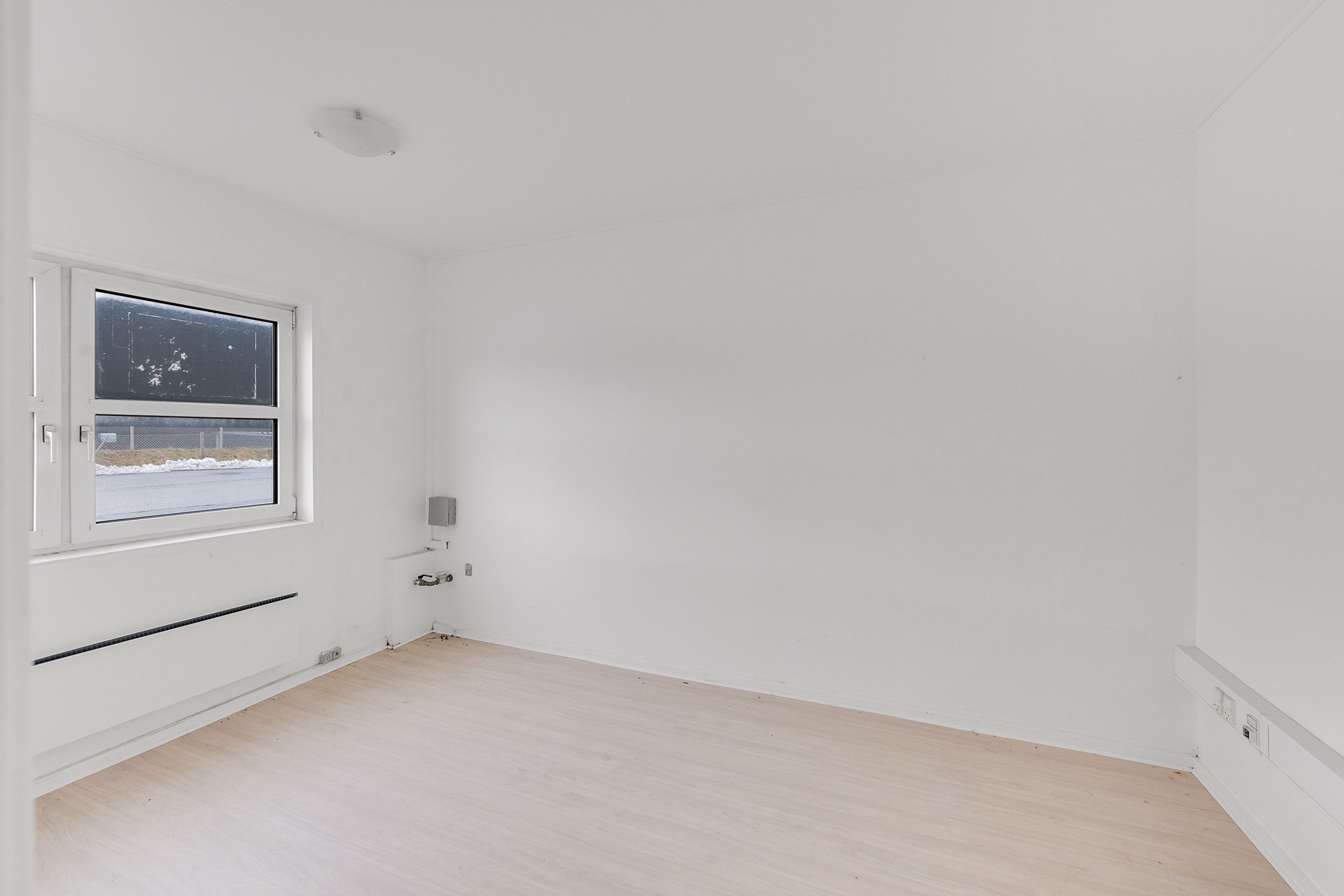 Et rent, tomt værelse med hvide vægge, lysegul trægulv og et vindue med dobbeltruder og udsigt til en sneduftet vej