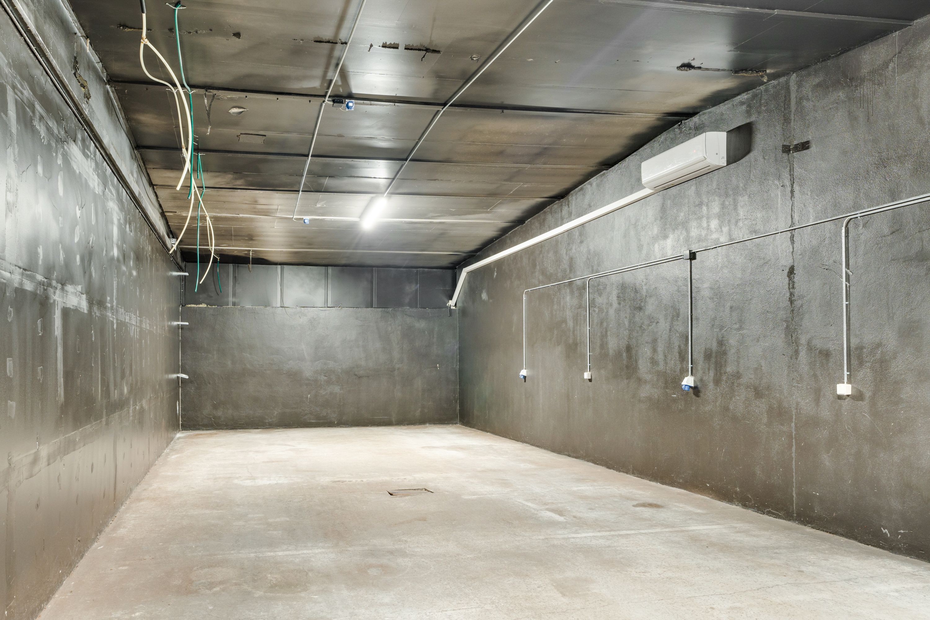 Et stort, tomt rum med lysegrå vægge og cementgulv, hvor der er installeret klimaanlæg og ledningsforløb