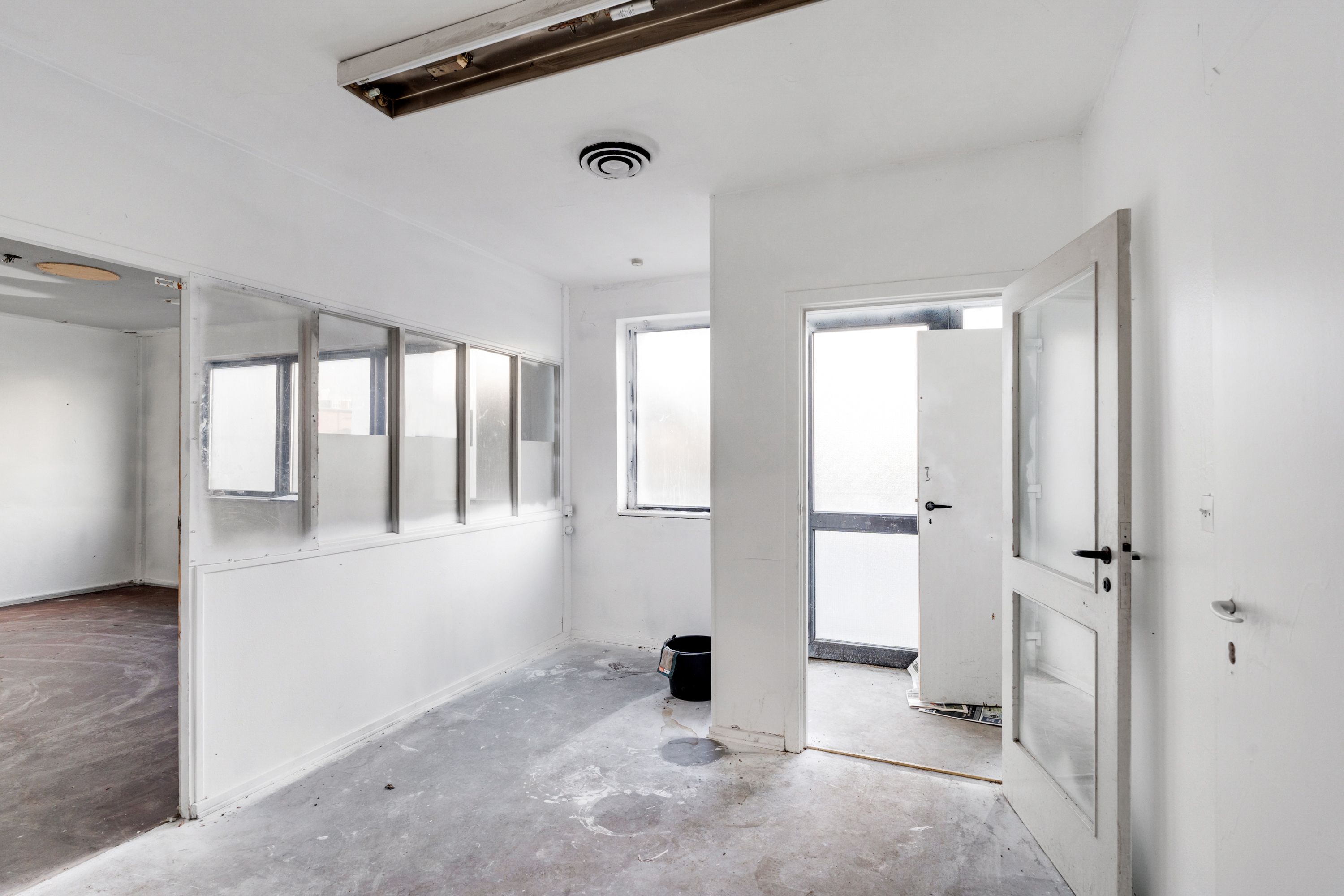 Hjørnerum med åbne døre, flere vinduer og en hvid væg, gulv og loft, samt en bøtte ved en dør