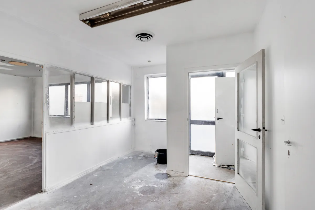 Hjørnerum med åbne døre, flere vinduer og en hvid væg, gulv og loft, samt en bøtte ved en dør