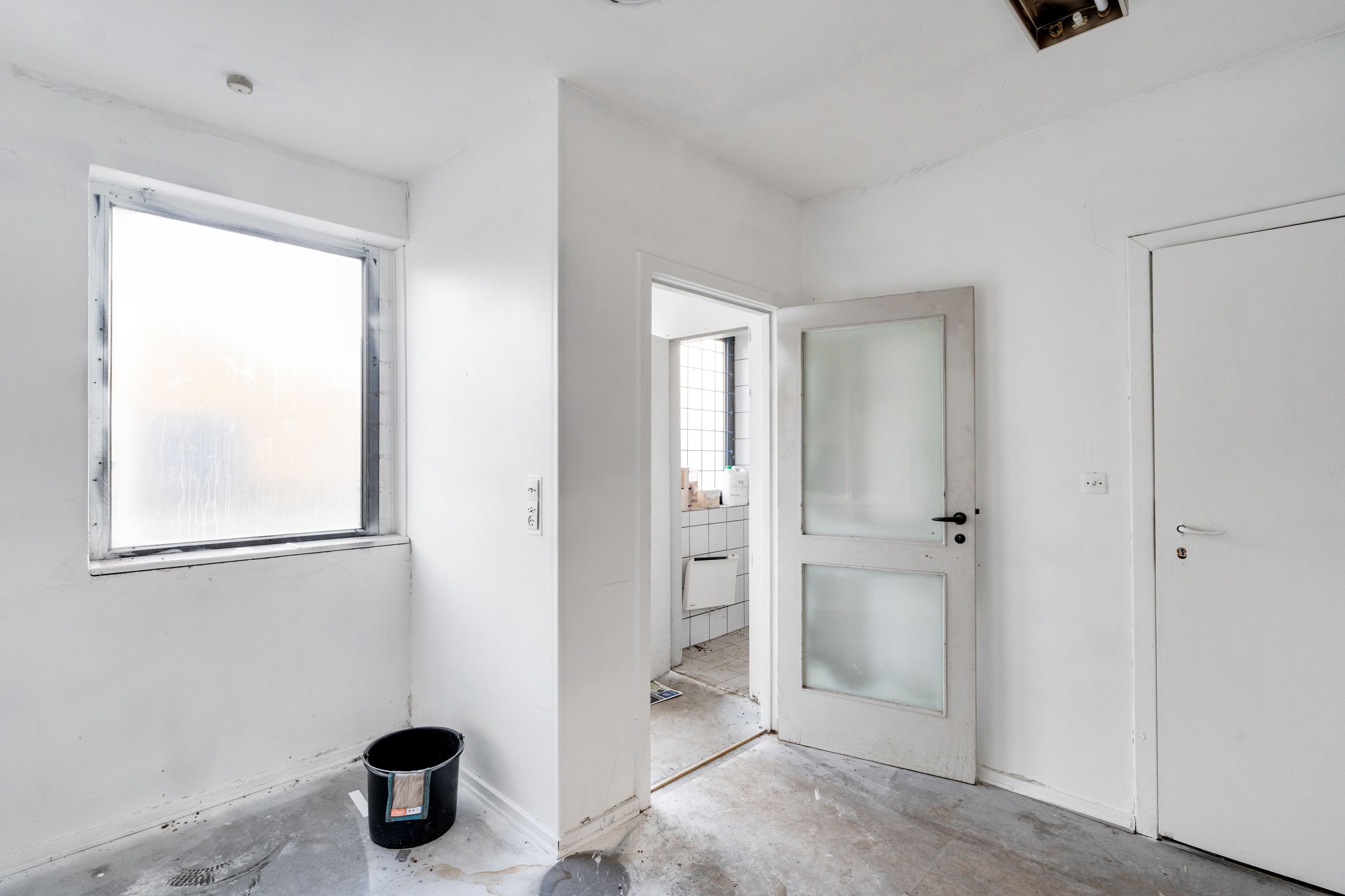 En tom rum med hvide vægge, gulv og loft. To hvide døre er åbne. Et skraldspand er placeret på gulvet.