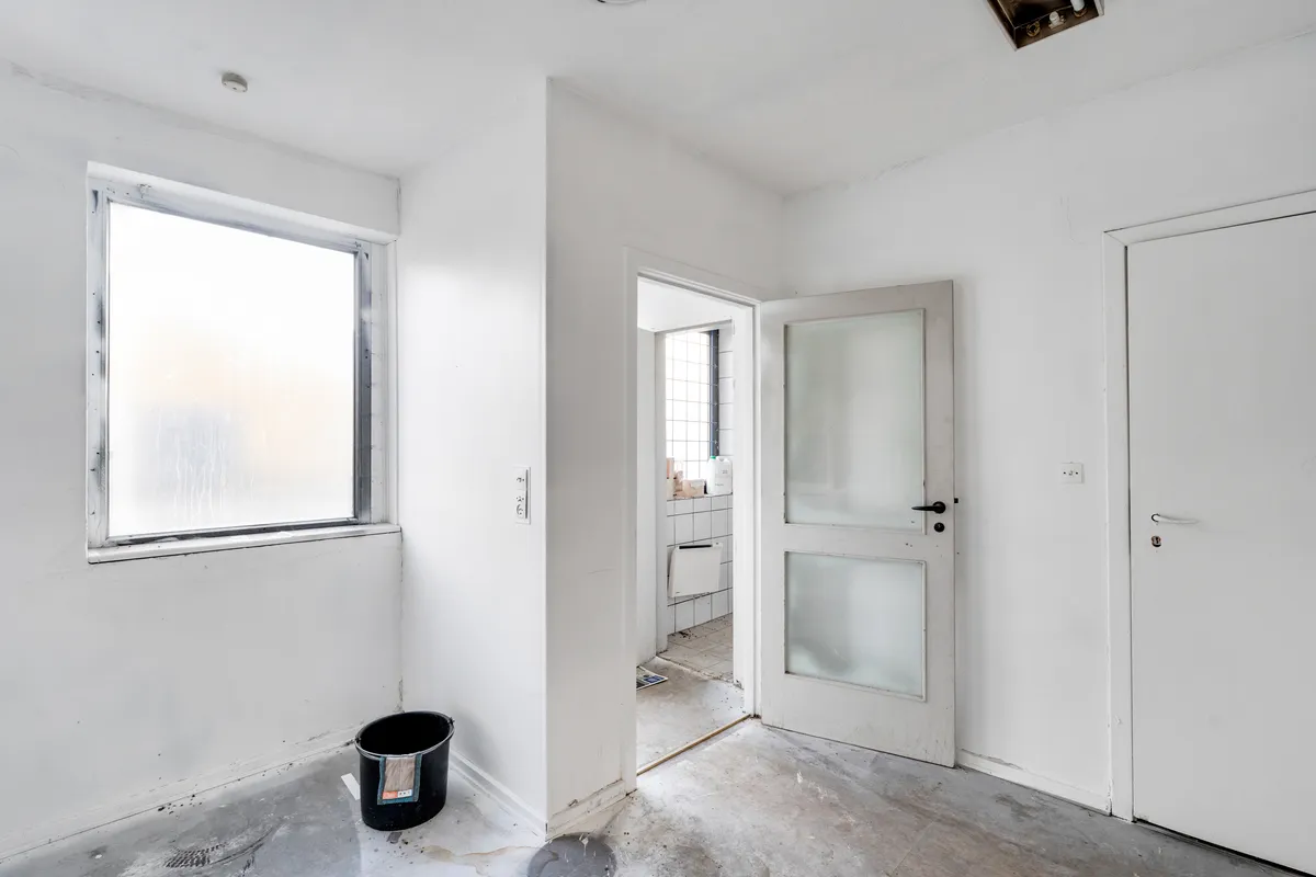 En tom rum med hvide vægge, gulv og loft. To hvide døre er åbne. Et skraldspand er placeret på gulvet.