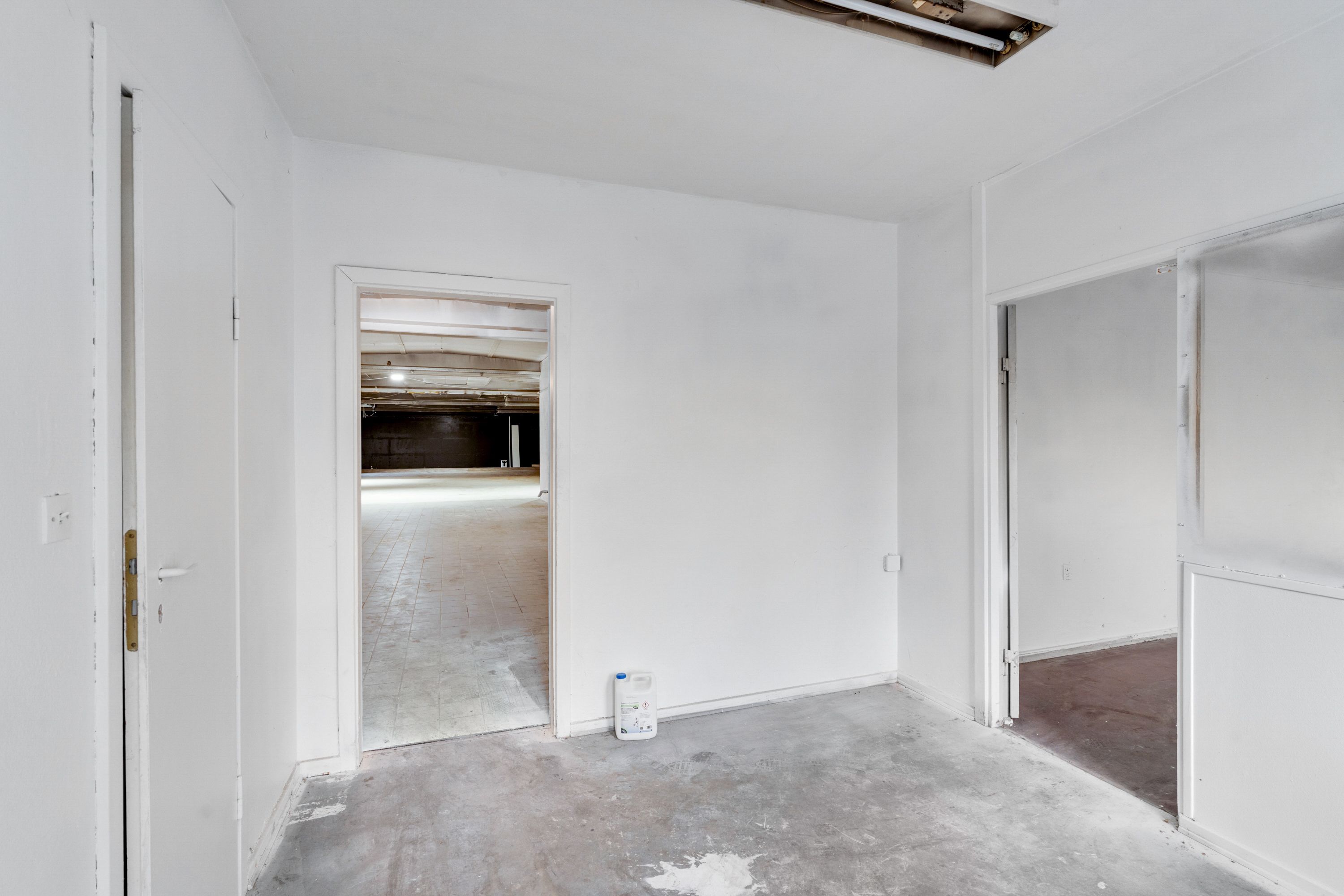 Et tomt lagerrum med to døre og en dør der fører ned til en garage, med en flaske på gulvet og en åben dør til højre