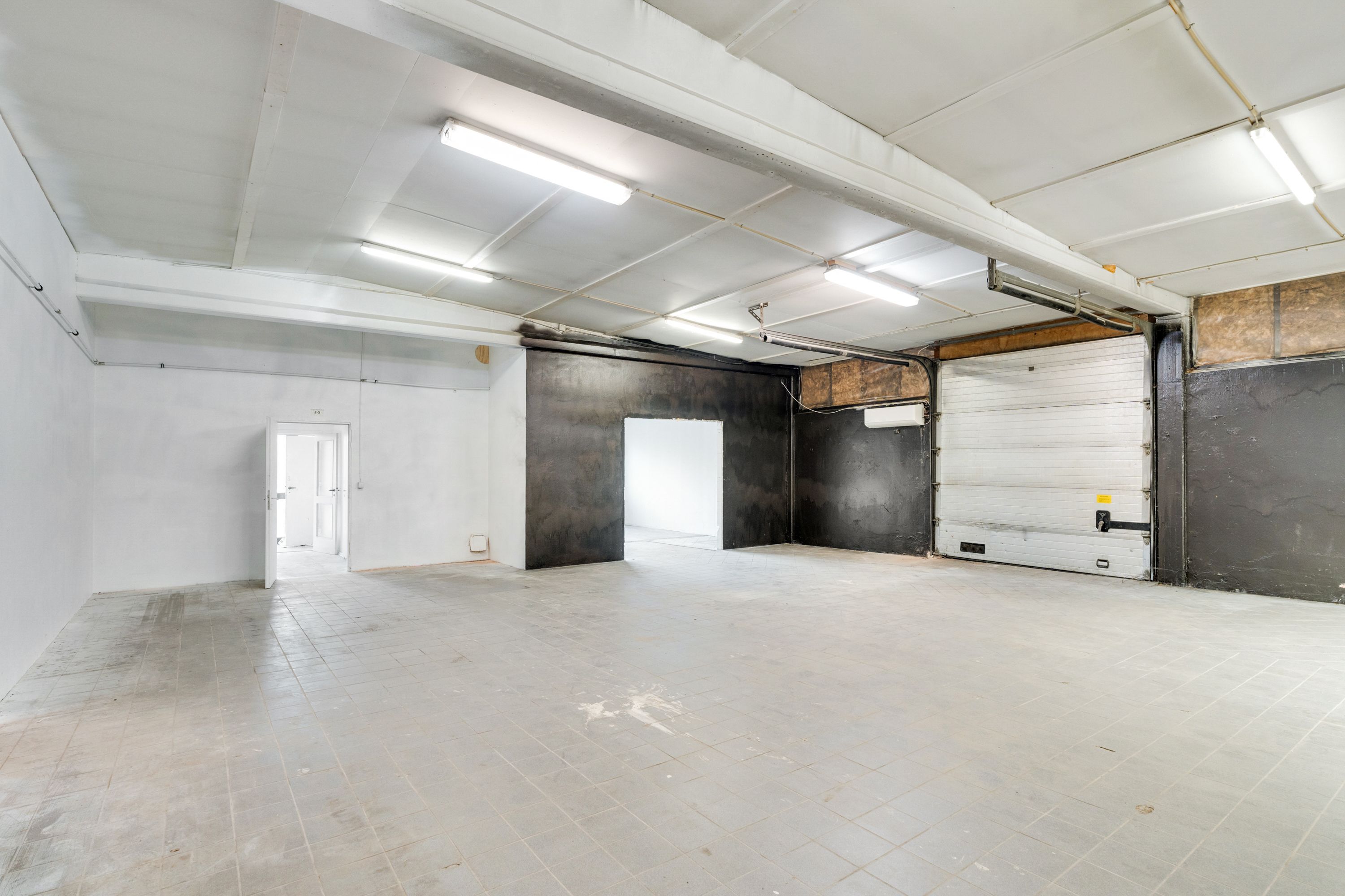 Et stort tomt rum med hvide vægge og hvidgule loftsbelægninger, to garageporte og en åben dør