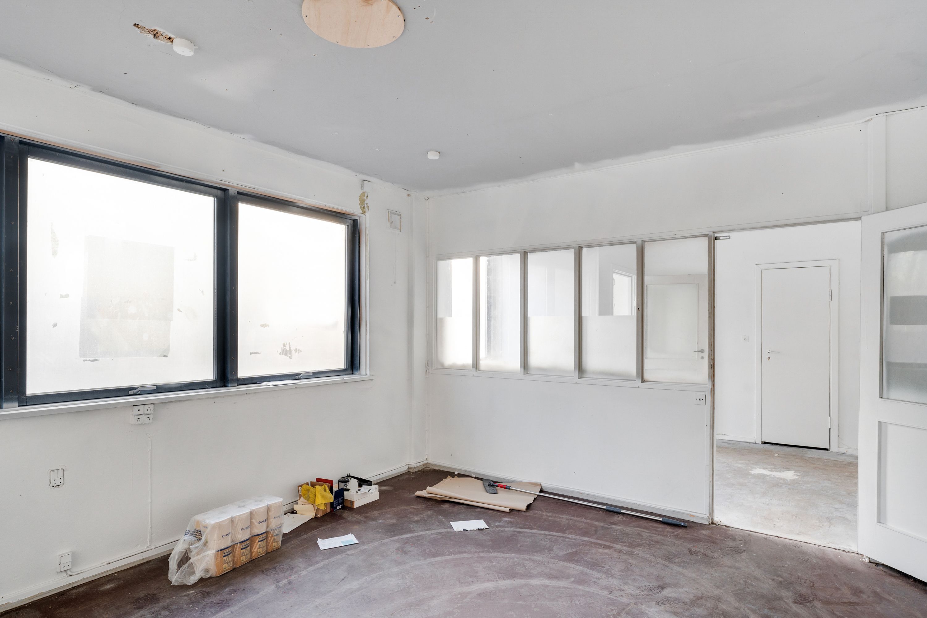 Et stort værelse med vinduer og to døre, der er lige blevet malet hvide. Et bord og flere stel med værktøj ligger på gulvet