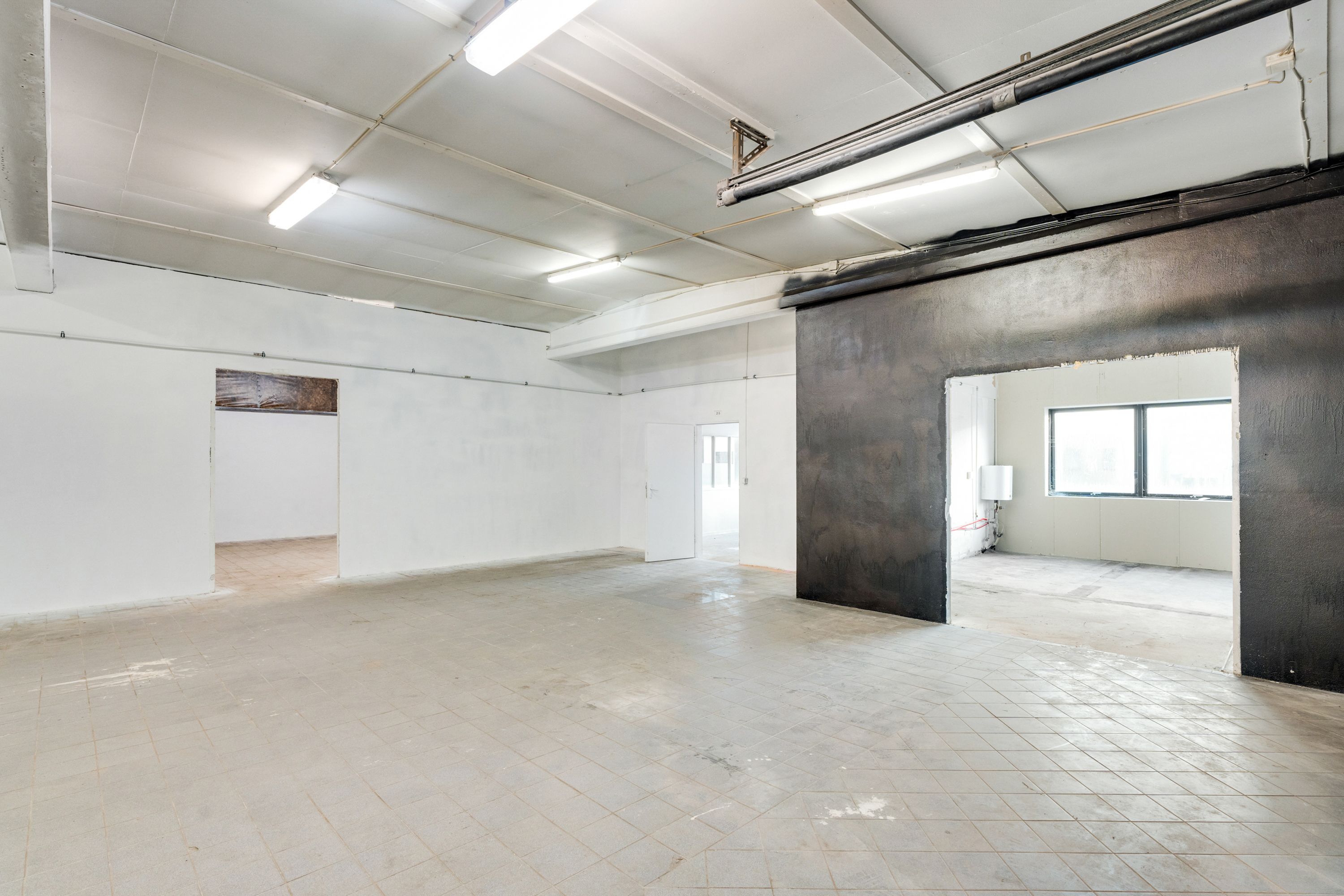 Lager med kontorplads til leje, inde med hvide vægge og store åbninger mod rummets udkant. Lamper hængende i loftet og