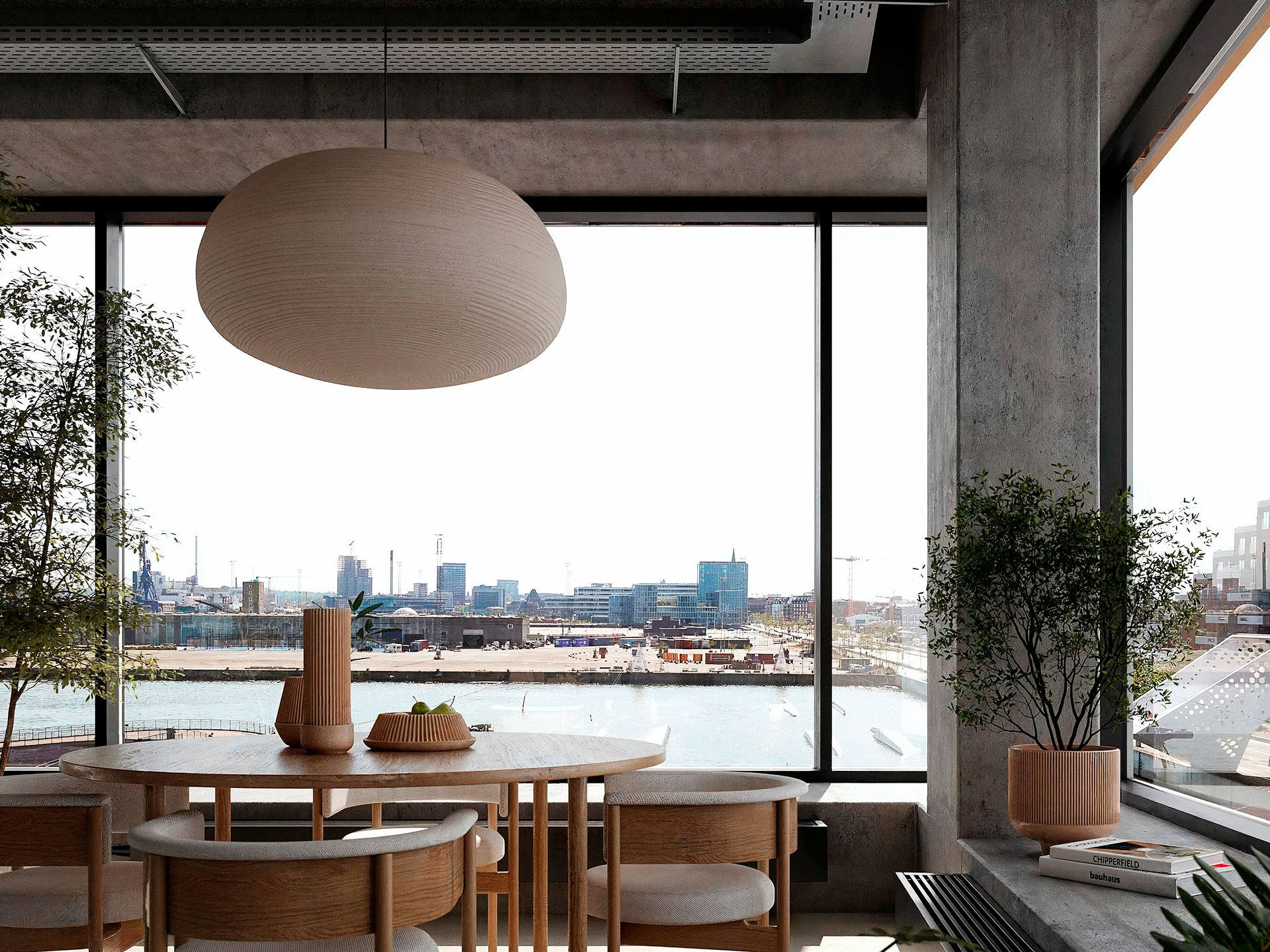 Vindue med udsigt til byens skyline og vand samt et rundt loftslys og træbord med en skål frugt og en vase