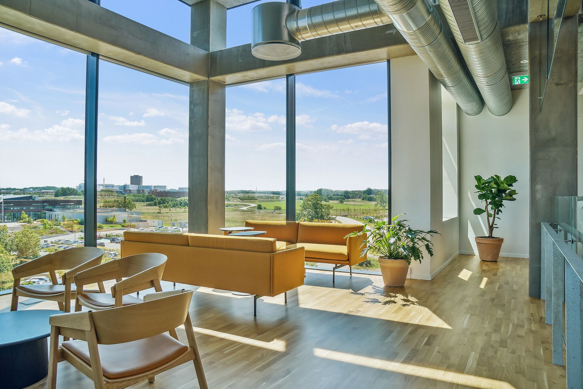 En lobby med møbler og plante i en bygning med store ruder, der giver udsigt til et landskab med grønne marker og bygninger