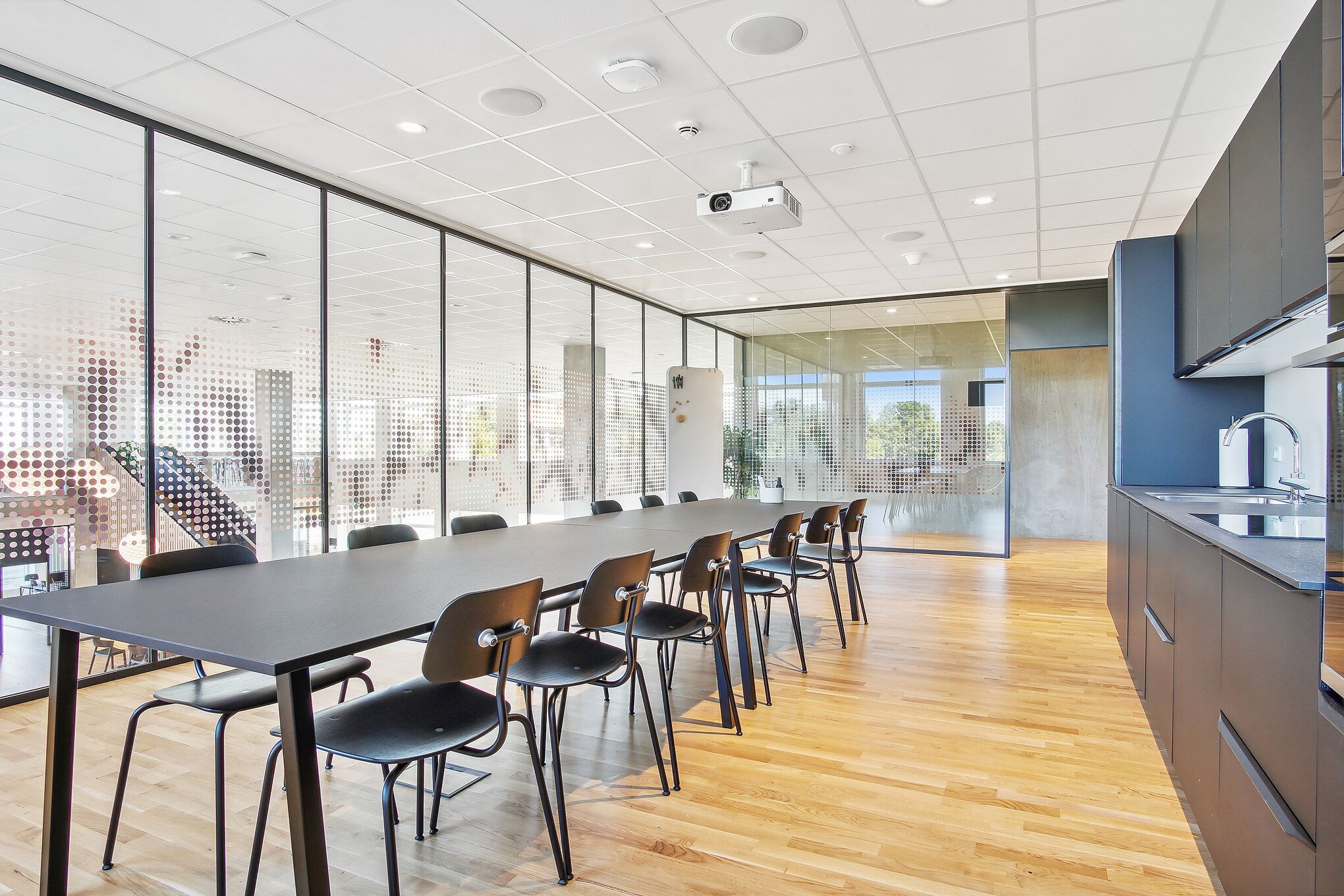 En stor, mørk konferencebord med mange stole er placeret i et rum med glasvægge, lyse loft og masser af lys.