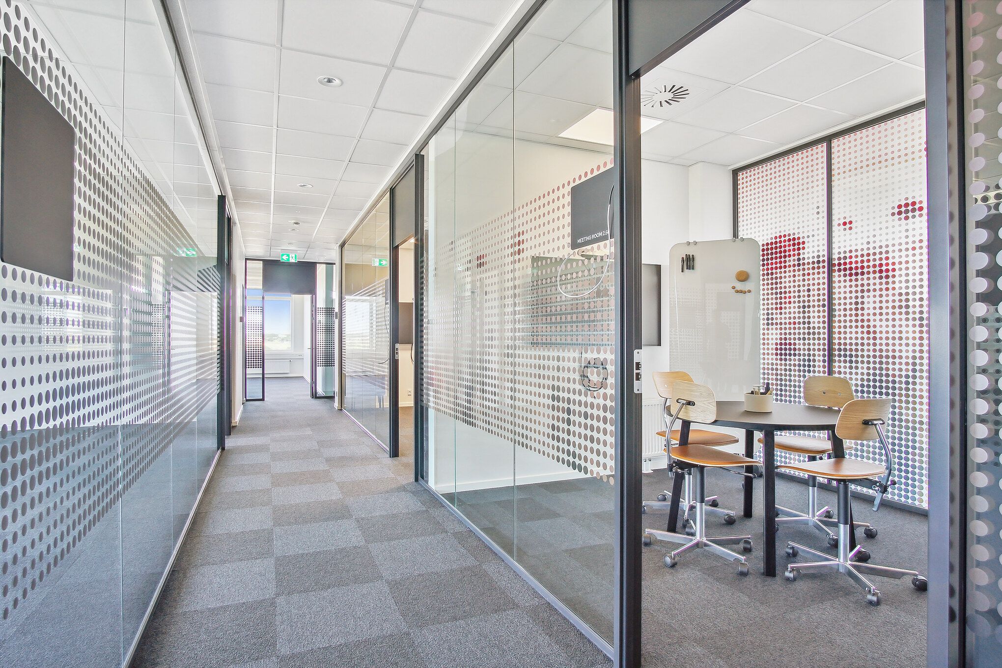 En moderne mødelokation med glas og metalvægge, to runde borde, fire stole og et hvidt køleskab med et lyserødt mønster på
