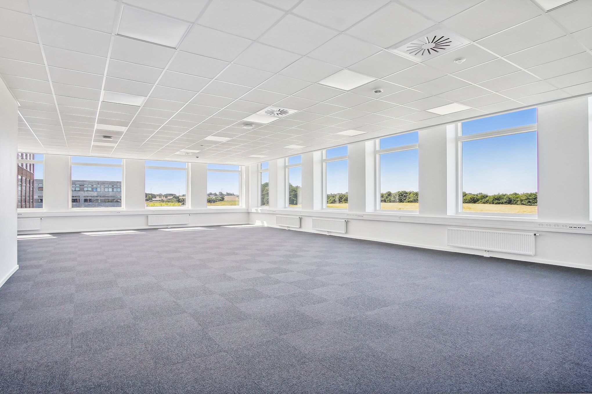 Et tomt kontor med store vinduer og en grøn have udenfor, hvor solen skinner og reflekterer i vinduerne