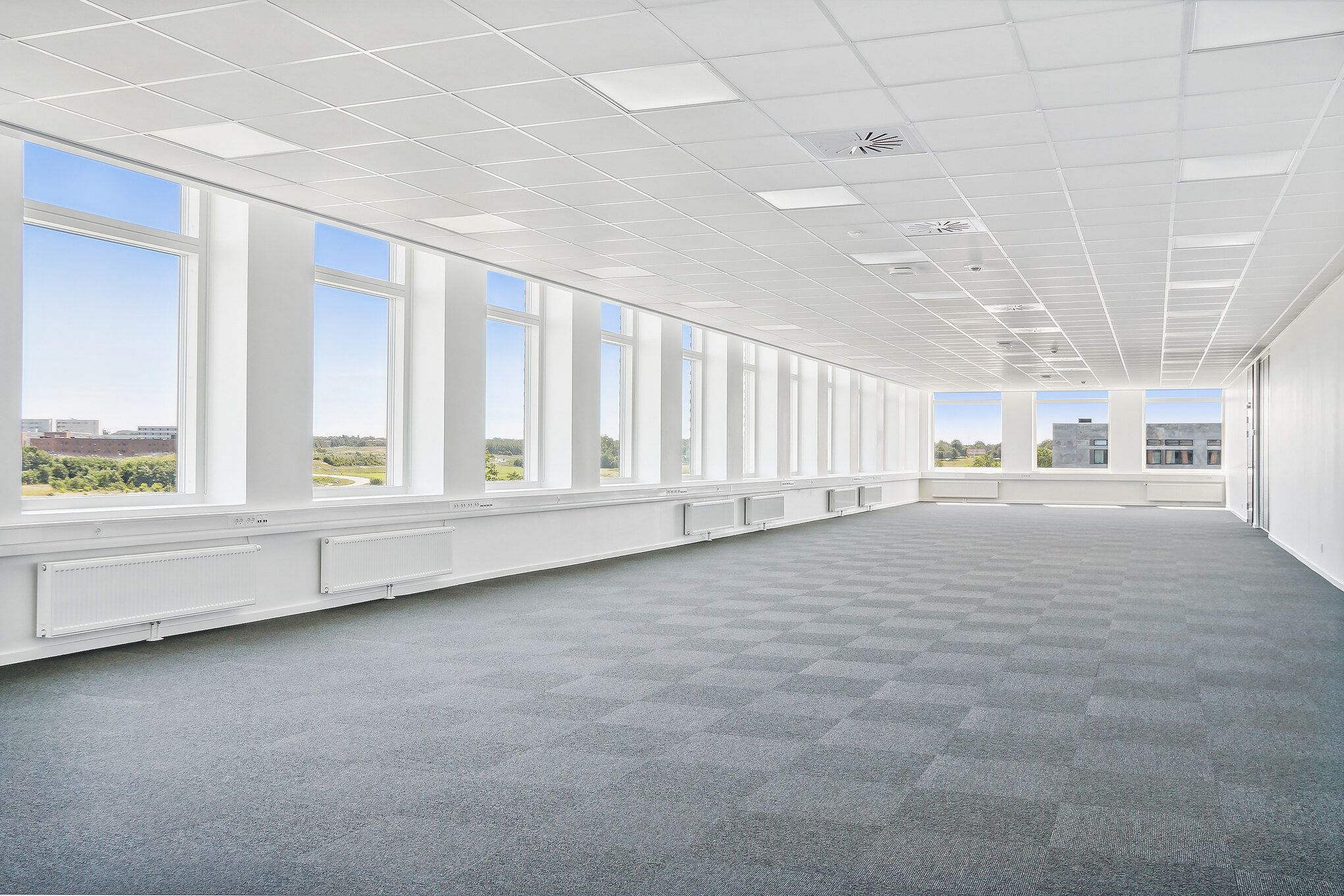 Brede kontorfællesskaber med store vinduer og hvide lofter samt radiatorer og lysegrå gulve