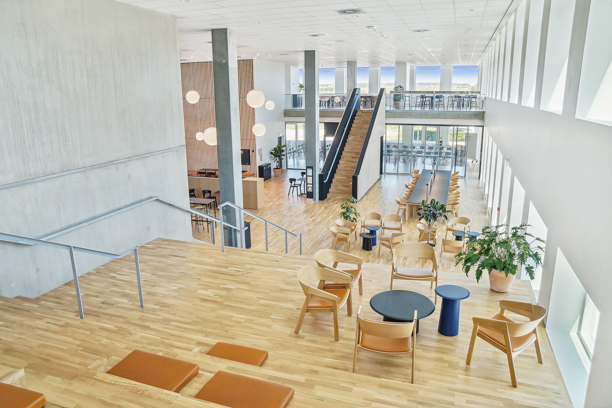 Et moderne lobbyområde med høje lofter, lyse gulve og smukke møbler. Her er flere træstole og -borde, og der er en terrasse