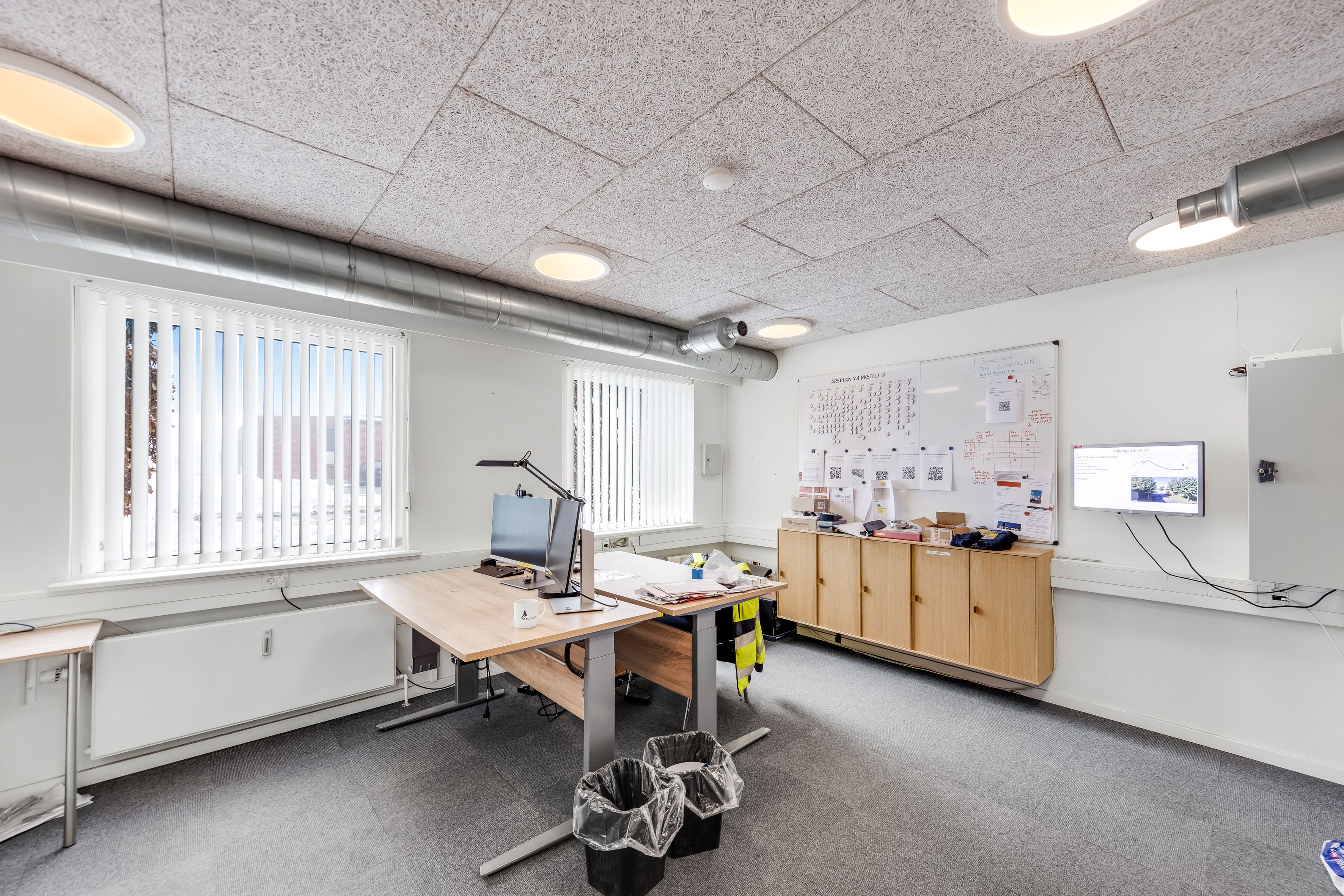 Ledig og funktionelt kontorlokal med skrivebord, computer, højloftet og lyse lysekroner samt ventilationsrør.