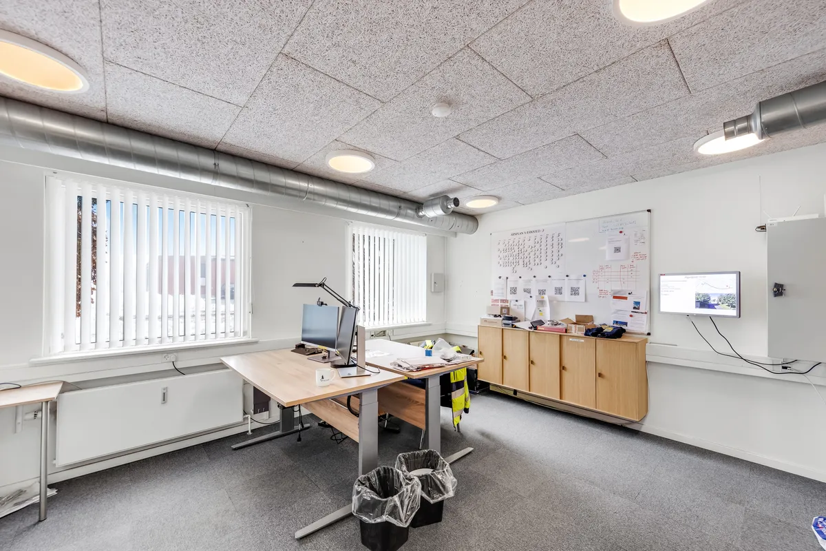 Ledig og funktionelt kontorlokal med skrivebord, computer, højloftet og lyse lysekroner samt ventilationsrør.