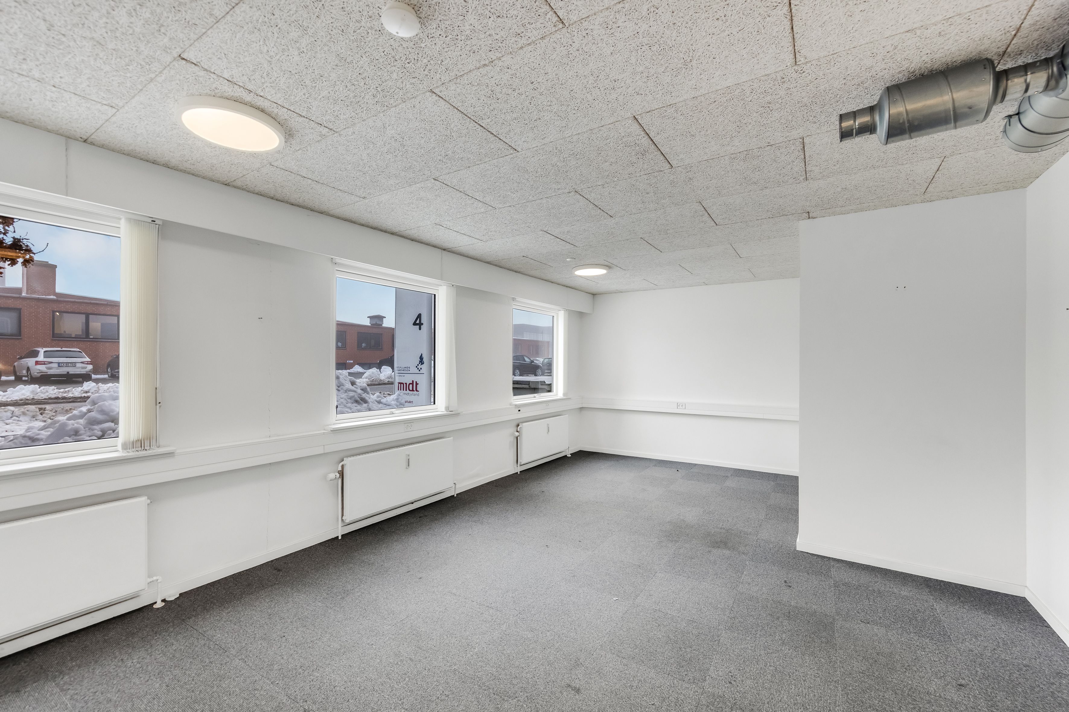 Ledig og rummelig lagerhal med højloft, tre vinduer, hvide vægge, radiatorer og gulvbelægning af gråt tæppe og nummeret fire