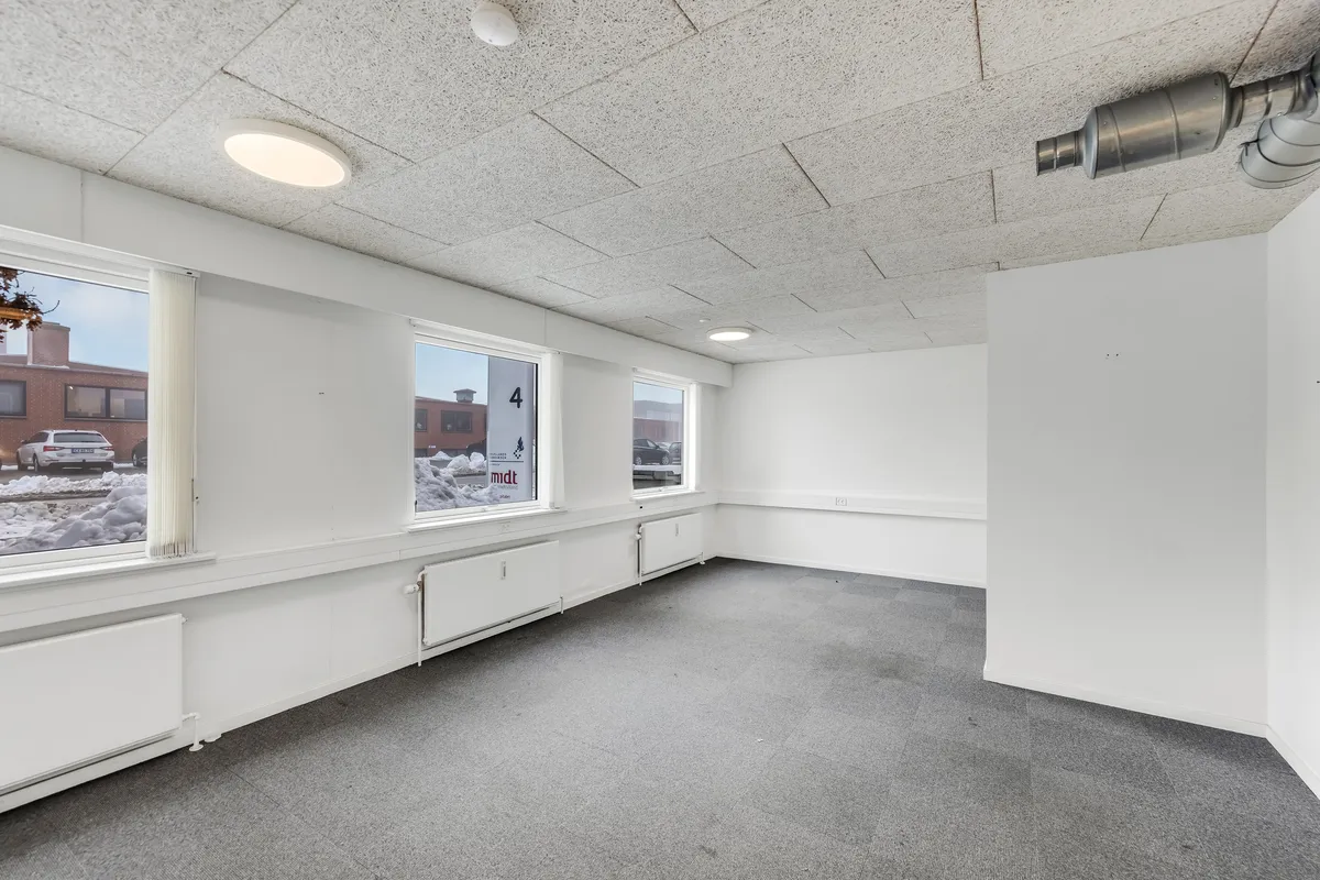 Ledig og rummelig lagerhal med højloft, tre vinduer, hvide vægge, radiatorer og gulvbelægning af gråt tæppe og nummeret fire